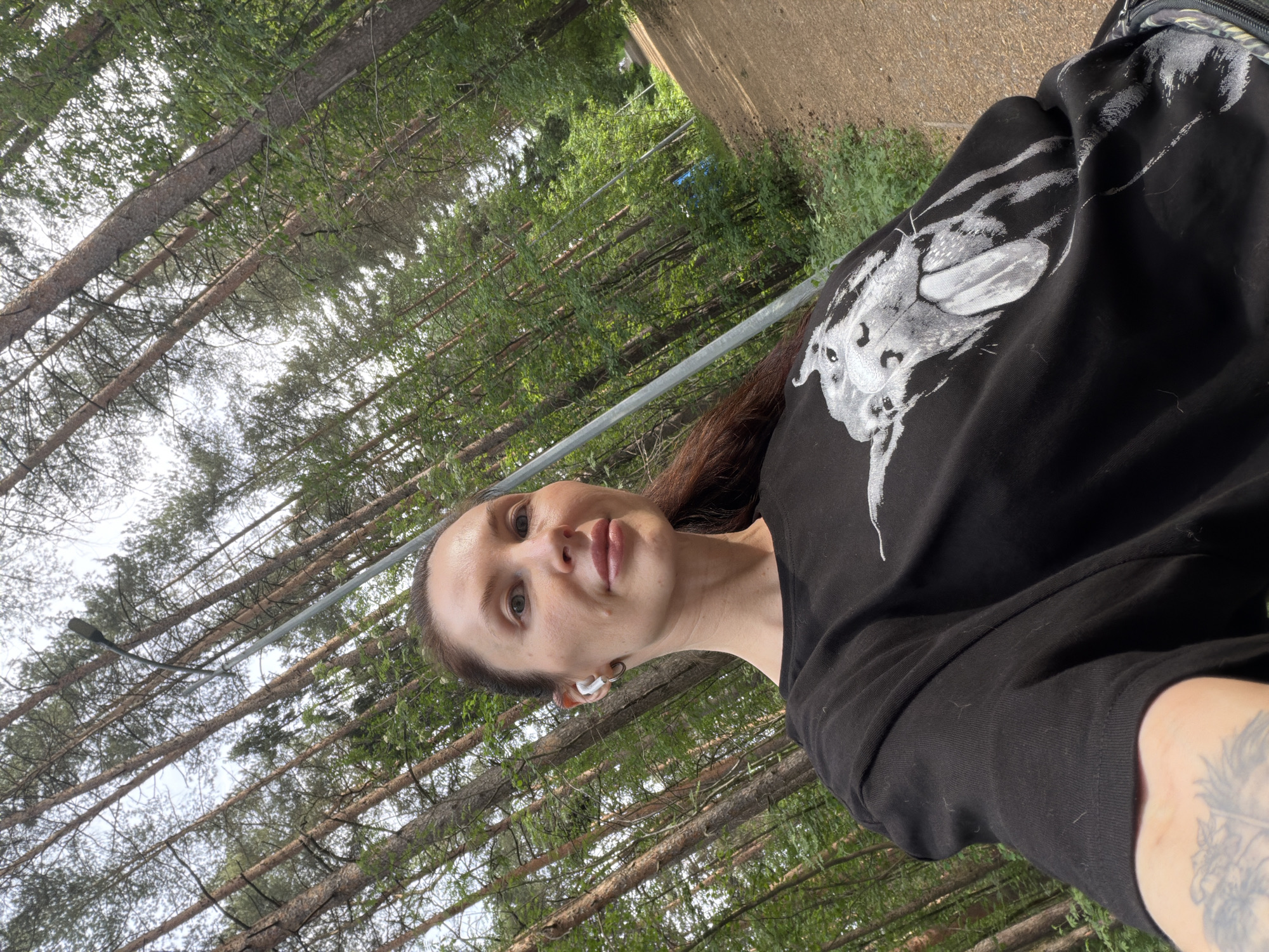 A woman is holding the camera up to take a selfie while standing outdoors. She is wearing a black sweatshirt with a white graphic design on the front. She has dark hair and is wearing a white earbud in her left ear. A tattoo is visible on her left arm. The background consists of a dense forest of tall trees. A gray, slightly rusty steel cable is visible diagonally across the center of the frame. A concrete structure and road are partially visible in the top right corner. The photo was likely taken in the city of Perm, Russia.