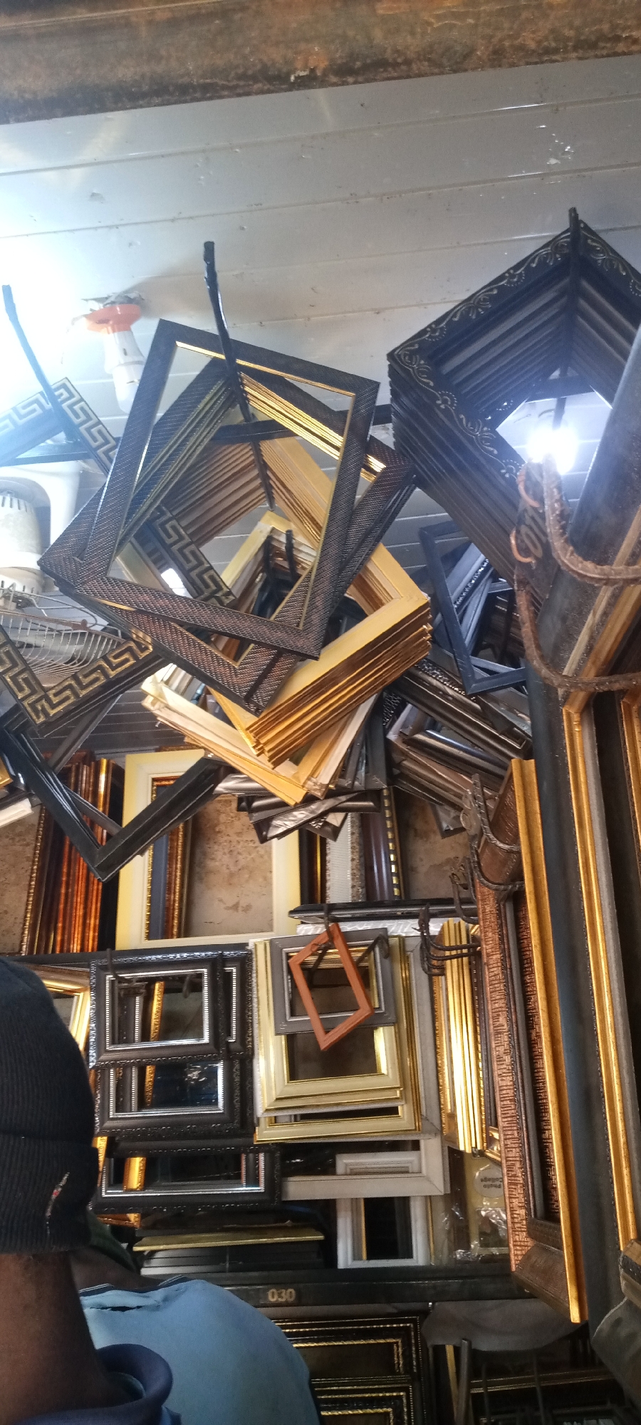The image shows a shop or workshop in Jos, Nigeria, filled with various picture frames. Frames of different sizes, colors (primarily gold, black, and brown), and designs are stacked on shelves and hanging from the ceiling. A person wearing a black beanie and a blue shirt is partially visible in the foreground. The environment appears cluttered with a concentration of wooden picture frames.