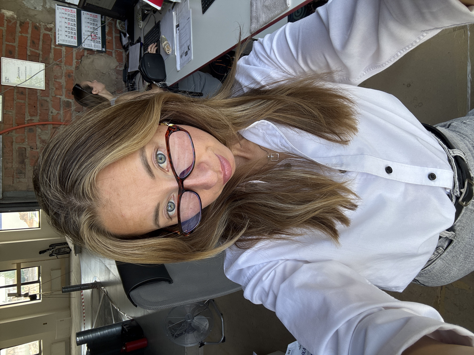 A young Caucasian woman is taking a selfie in an office environment. She has long blonde hair, is wearing glasses, and a white button-down shirt. She is seated in an office chair, with a desk and computer visible behind her. In the background, there are windows, a calendar, and another person in the office. The office appears to be a renovated industrial space, with exposed brick walls. The woman is the primary subject, and the overall tone of the image is casual and informal. There are no discernible elements that would indicate a specific location within Chaikovsky, Russia.
