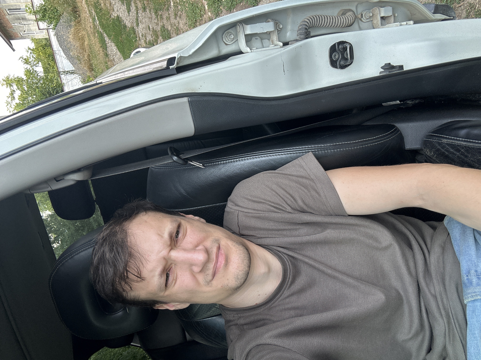 A man with short brown hair and a concerned expression is sitting in the driver's seat of a white car, with the passenger door open. He is wearing a brown t-shirt and blue jeans. The interior of the car is visible, showing black leather seats and a seatbelt. Outside the car, a dirt road is visible, along with trees, a fence, a pile of gravel, and a brick building. The scene appears to be in a rural or suburban area. The context suggests the photo was taken in Makiv, Ukraine.