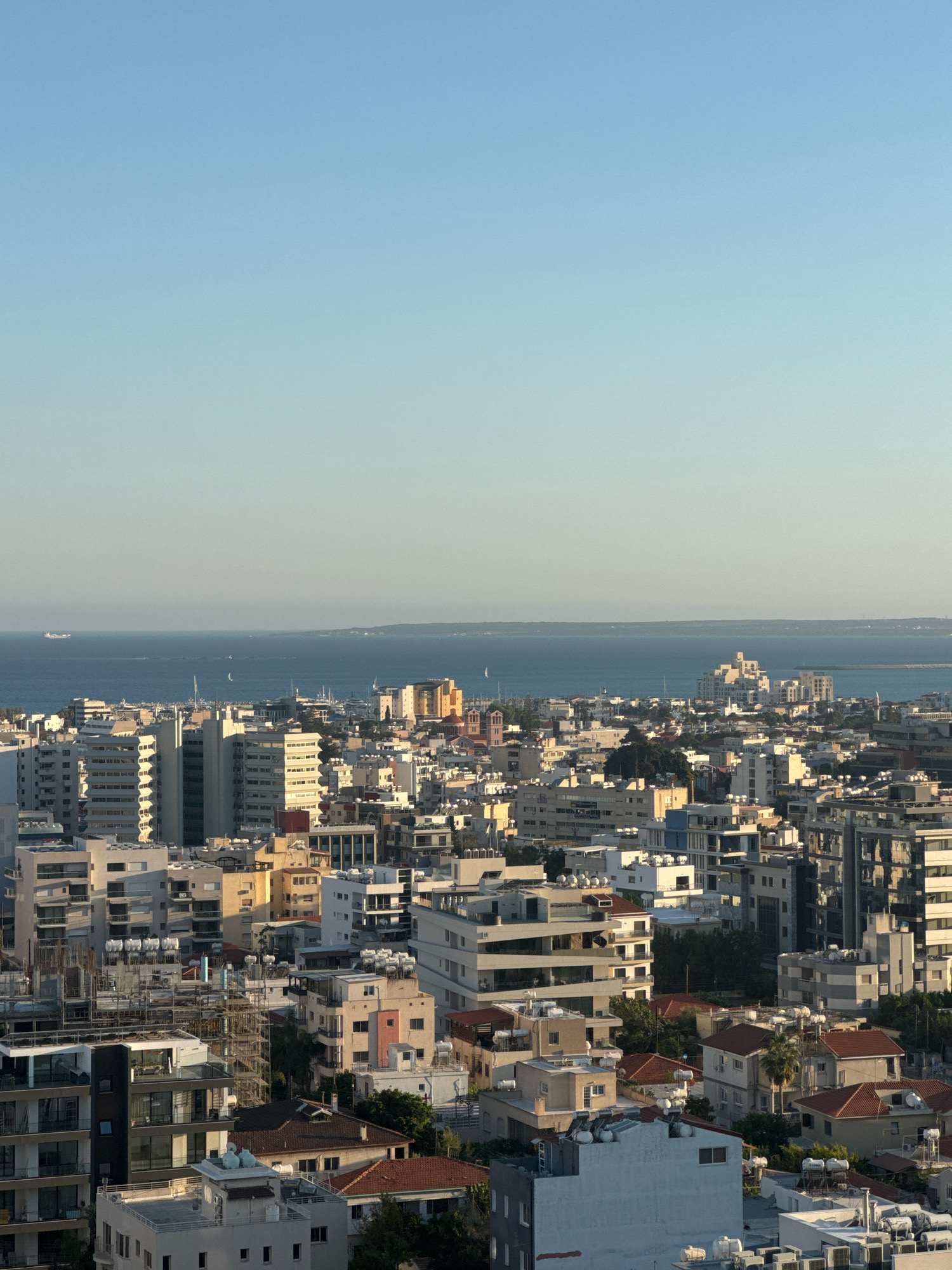 This is a high-angle, full-frame, eye-level shot of a densely populated urban area bordering a body of water, likely the Mediterranean Sea, under a clear, pale blue sky. The scene captures the coastal city of Limassol, Cyprus, with its characteristic mix of residential and commercial buildings.
The foreground is dominated by a collection of multi-story buildings, varying in architectural style and age. Many of these structures are apartment blocks with balconies, while others appear to be commercial spaces. Some buildings are under construction, indicated by scaffolding and exposed concrete frameworks. Rooftops are visible, many equipped with solar water heaters.
Further back, the urban landscape continues, with a higher density of buildings. The architecture displays a range of colors, predominantly white, cream, and beige, with accents of yellow and grey. Some buildings have a more modern aesthetic with glass facades, while others are older and more traditional. Trees are interspersed among the buildings, providing a touch of greenery.
In the mid-ground and background, the city extends towards the sea. The water is a deep blue, reflecting the sky. Several boats, including sailboats and a larger cargo ship, are visible on the water, suggesting active maritime traffic. The horizon is marked by a low-lying landmass in the distance, indicating the presence of other coastal features or islands.
The lighting suggests it is either early morning or late afternoon, with soft, directional sunlight casting shadows across the buildings. There are no people or vehicles clearly discernible in the image. The overall impression is one of a bustling coastal city at peace.