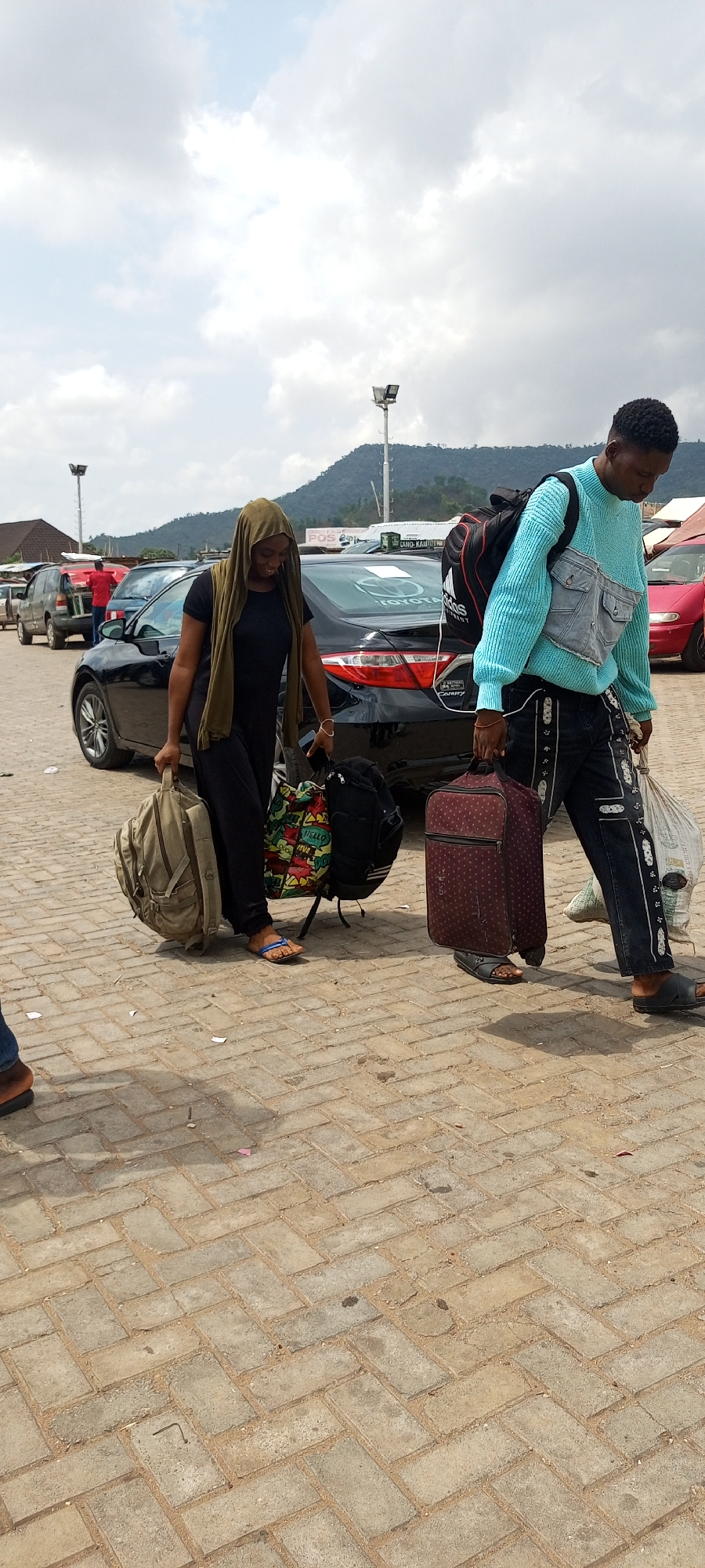The image shows a man and a woman walking across a paved lot, carrying luggage. The woman is wearing a dark-colored dress and a tan headscarf. She is carrying a tan backpack, and what appears to be a colorful bag. The man is wearing a blue sweater, jeans, and sandals. He has a black backpack on and is carrying a red suitcase and a plastic bag.
Behind them are several parked cars, including a black Toyota Camry. In the background, there is a mountain range. A sign is visible behind the car; it may say "POS". The location is indicated as the city of Lokoja, Nigeria, but there is no visual verification of this location.