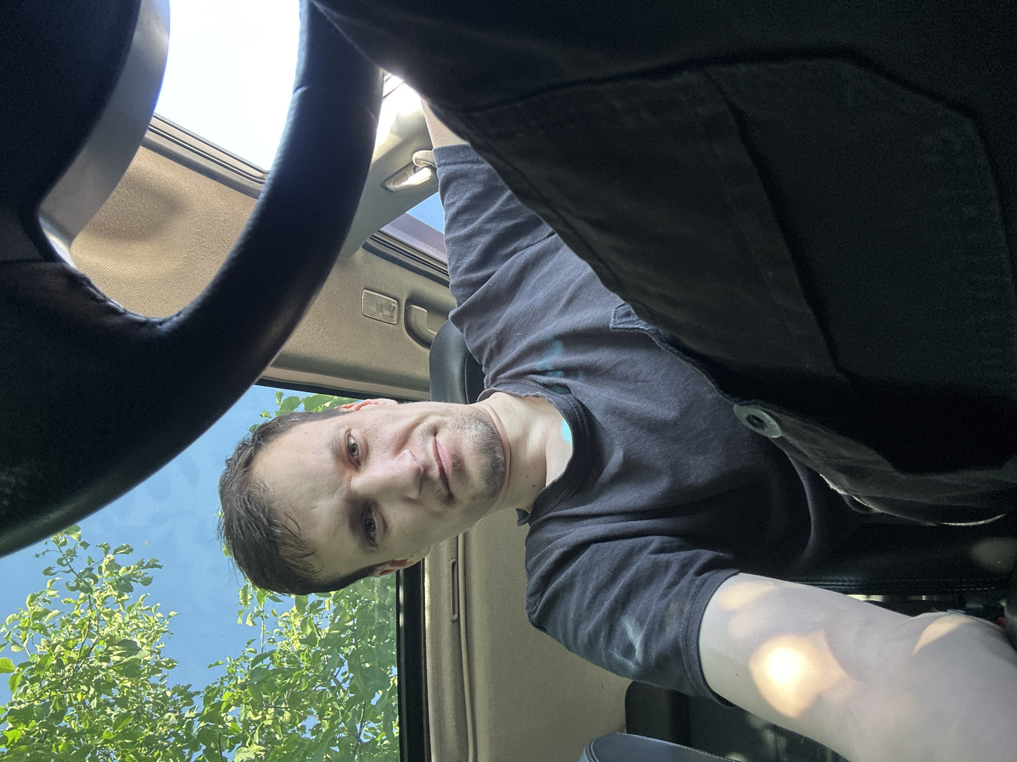 This is a selfie taken from inside a car. A man with short brown hair and a light beard is looking at the camera. He is wearing a dark t-shirt and dark pants. The sun is shining through an open sunroof, casting a bright light on his left arm. Green leaves and blue sky are visible through the sunroof. The steering wheel of the car is in the foreground. The image does not provide any specific information about the location in the city of Shatava, Ukraine.