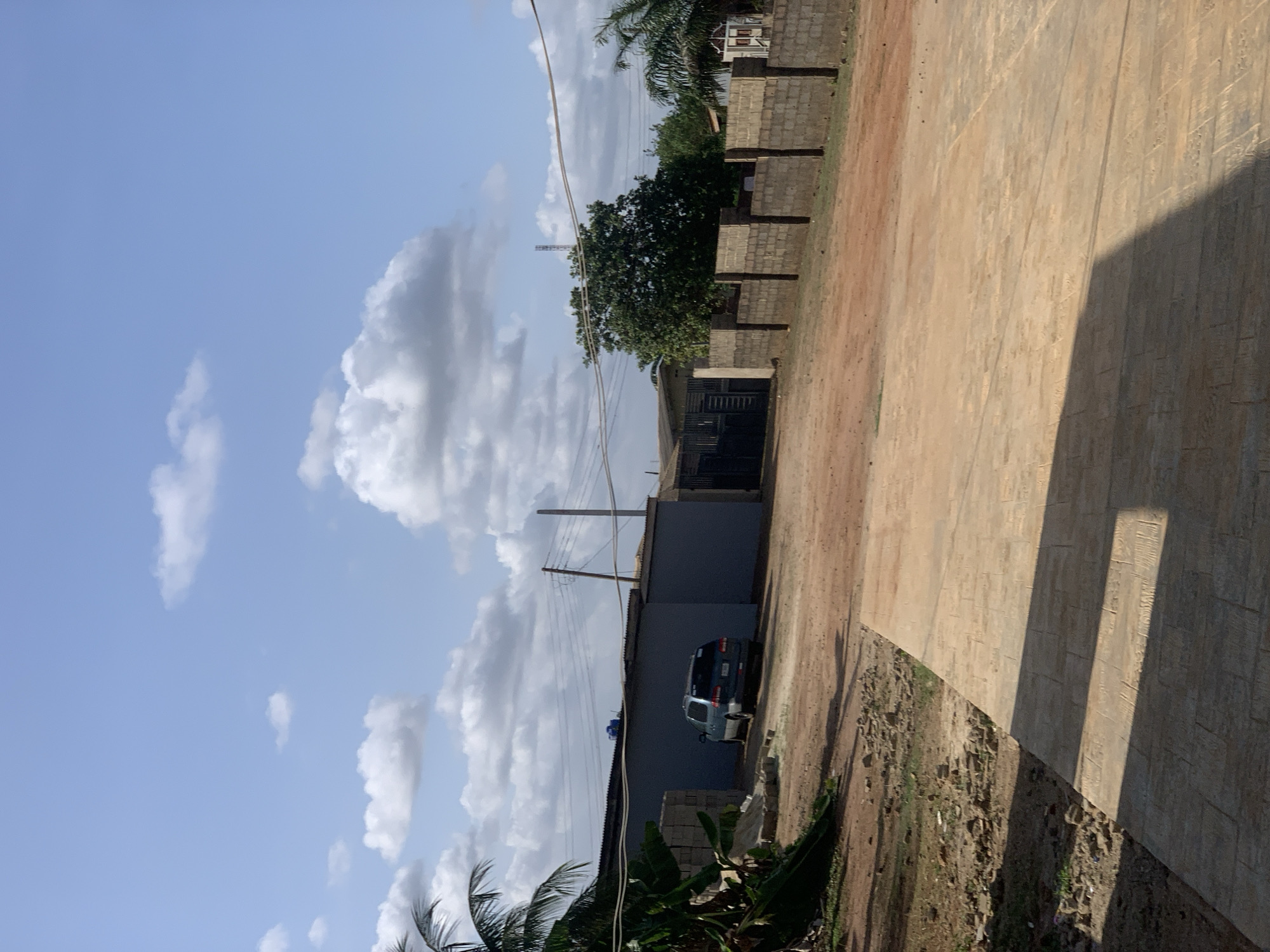 An outdoor scene in Ikorodu, Nigeria, under a partly cloudy sky. A dark-colored SUV is parked next to a gray, single-story building. A dirt road runs alongside the building, leading to a dark gate structure. A concrete block wall borders the dirt road on the far side. Palm trees and other vegetation are present in the foreground. A tall, slender communication tower is visible in the background. A large paved area with geometric patterns is prominent on the right side of the frame, casting a shadow. Electrical wires run overhead.