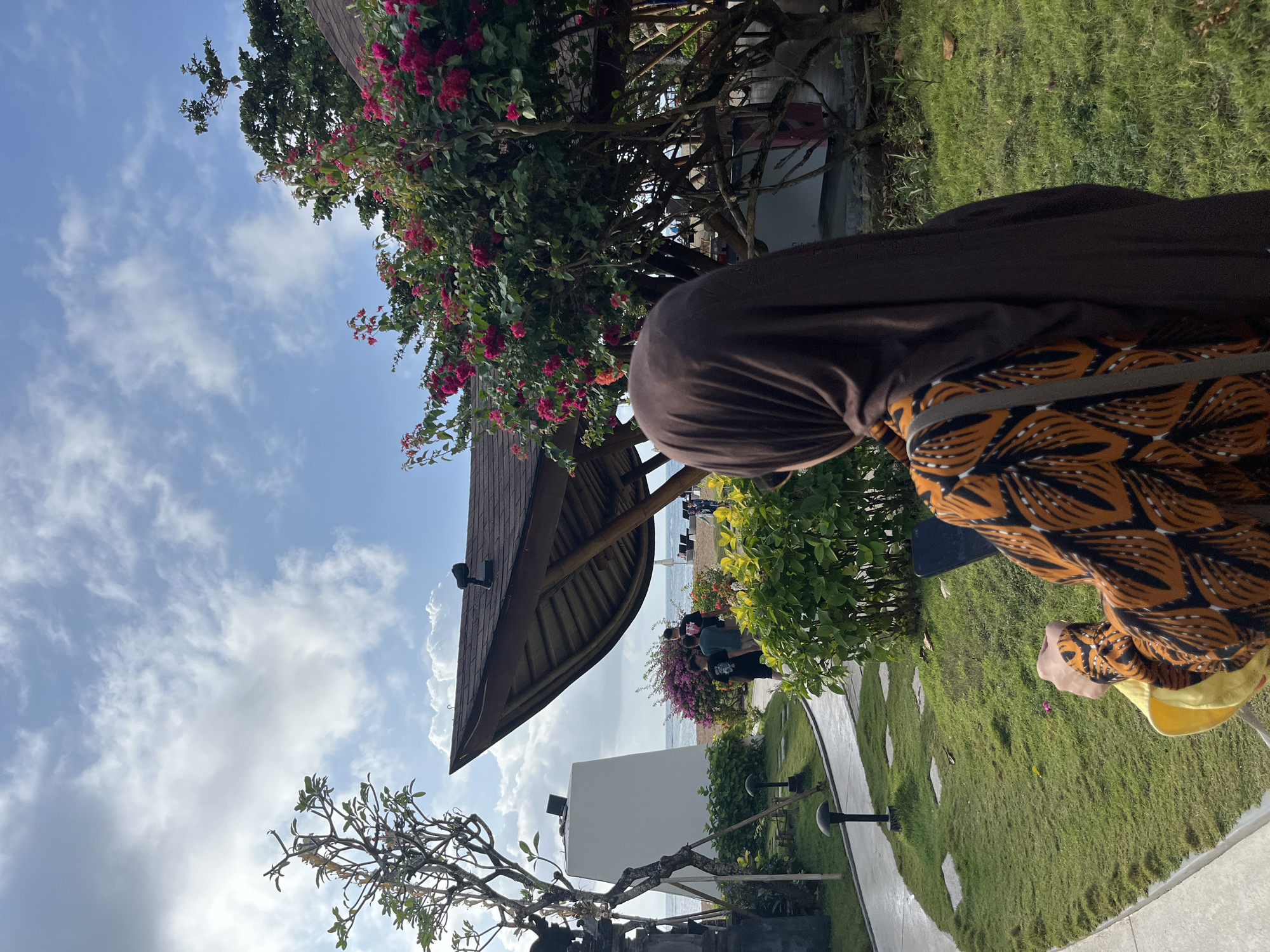 In Kuta Selatan, Indonesia, a woman wearing a brown hijab and an orange and black patterned dress is seen from behind, holding a mobile phone in her left hand. She is standing in a lush garden with green grass and pathways, adjacent to a beach with lounge chairs. A building with a distinctive curved roof is visible, and a tree with pink flowers grows nearby. In the background, several people are walking towards the beach under a sky with scattered clouds.
