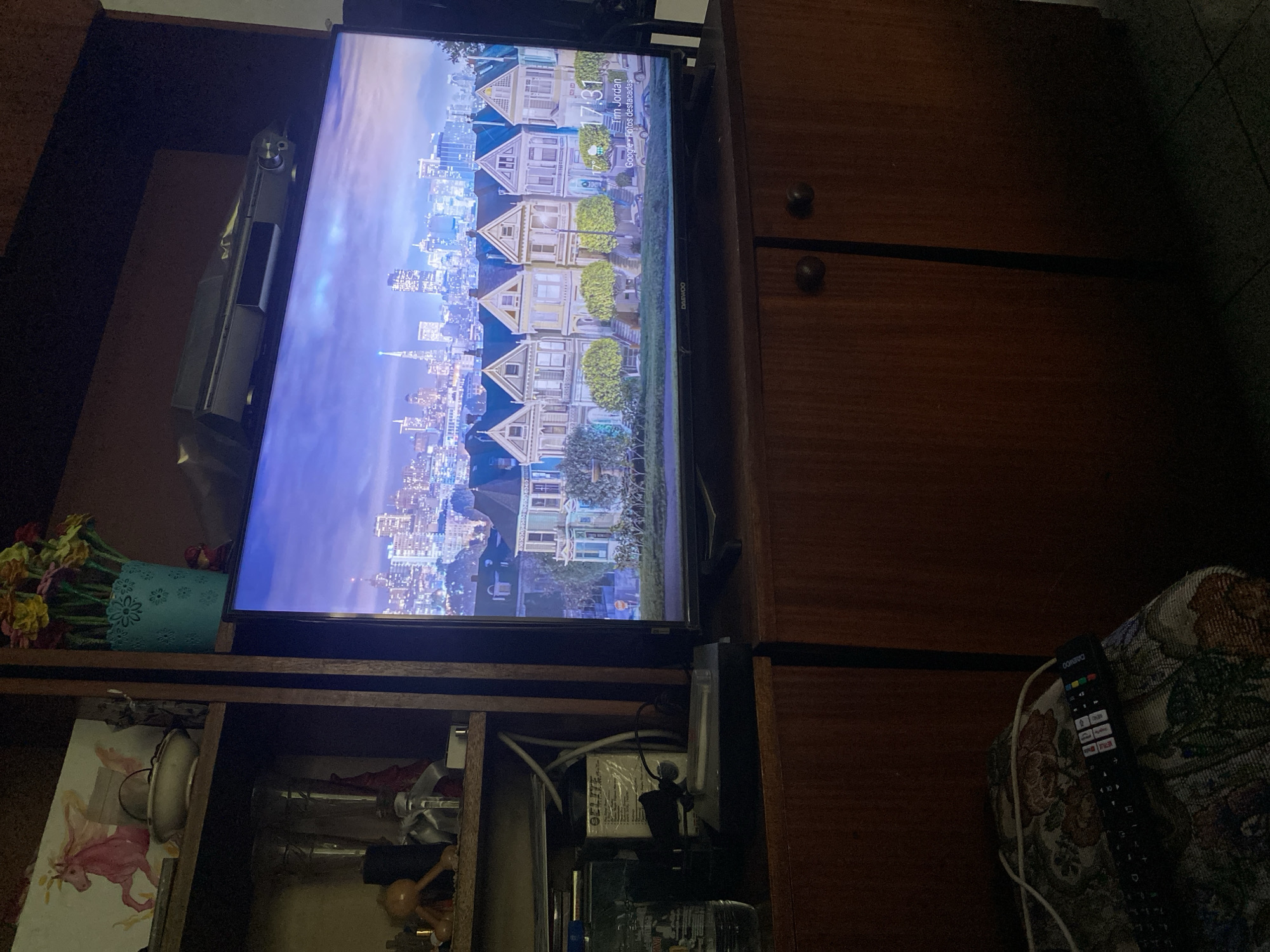The image displays a living room setting, likely within an apartment in Smolyan, Bulgaria, as indicated by the on-screen display of "Tim Jordan" and "Google - Fotos destacadas," which suggests a Google Photos screensaver. A large television screen, displaying a vibrant image of a city skyline with Victorian-style houses in the foreground, is the central focus. The screen is positioned above a wooden cabinet with two doors, each featuring a dark circular handle. To the left of the television, on a shelf within a dark wooden cabinet, a bottle of "GELIFE" is visible, along with various other unidentifiable items. Above the television, a metallic object, possibly a soundbar or media player, rests on the shelf. On the top shelf of the cabinet, a decorative arrangement of colorful artificial flowers in a blue pot is present, along with a pink painted image of a horse-like creature. In the lower portion of the image, a portion of a patterned sofa or chair with a remote control resting on it is visible. The overall lighting suggests an indoor, evening setting.