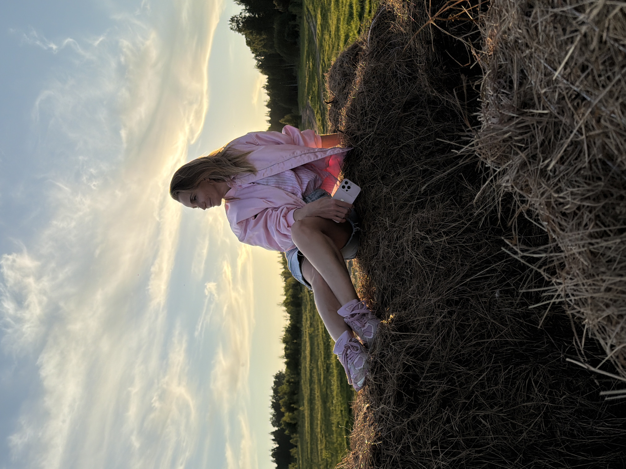 A young woman is captured sitting outdoors on a large, roughly textured hay bale. She is positioned with her body angled slightly towards the left of the frame, her head tilted downwards, and her gaze directed away from the camera. In her right hand, she holds a light-colored smartphone. She is dressed in a light pink jacket, a striped top beneath, denim shorts, and matching pink athletic shoes with white laces.
The scene unfolds during the golden hour, evident from the warm, soft light that illuminates the environment and casts subtle shadows. The sunlight gently backlights the wispy clouds in the sky and highlights the woman's hair. In the background, a verdant field stretches out, bordered by a dense line of trees or forest. The sky above is a clear, pale blue with scattered, delicate white clouds. The hay bale itself appears dry and composed of bundled straw. The overall mood is tranquil and natural. This outdoor setting is identified as Ozernoye-1, Russia. There is no visible text in the image.