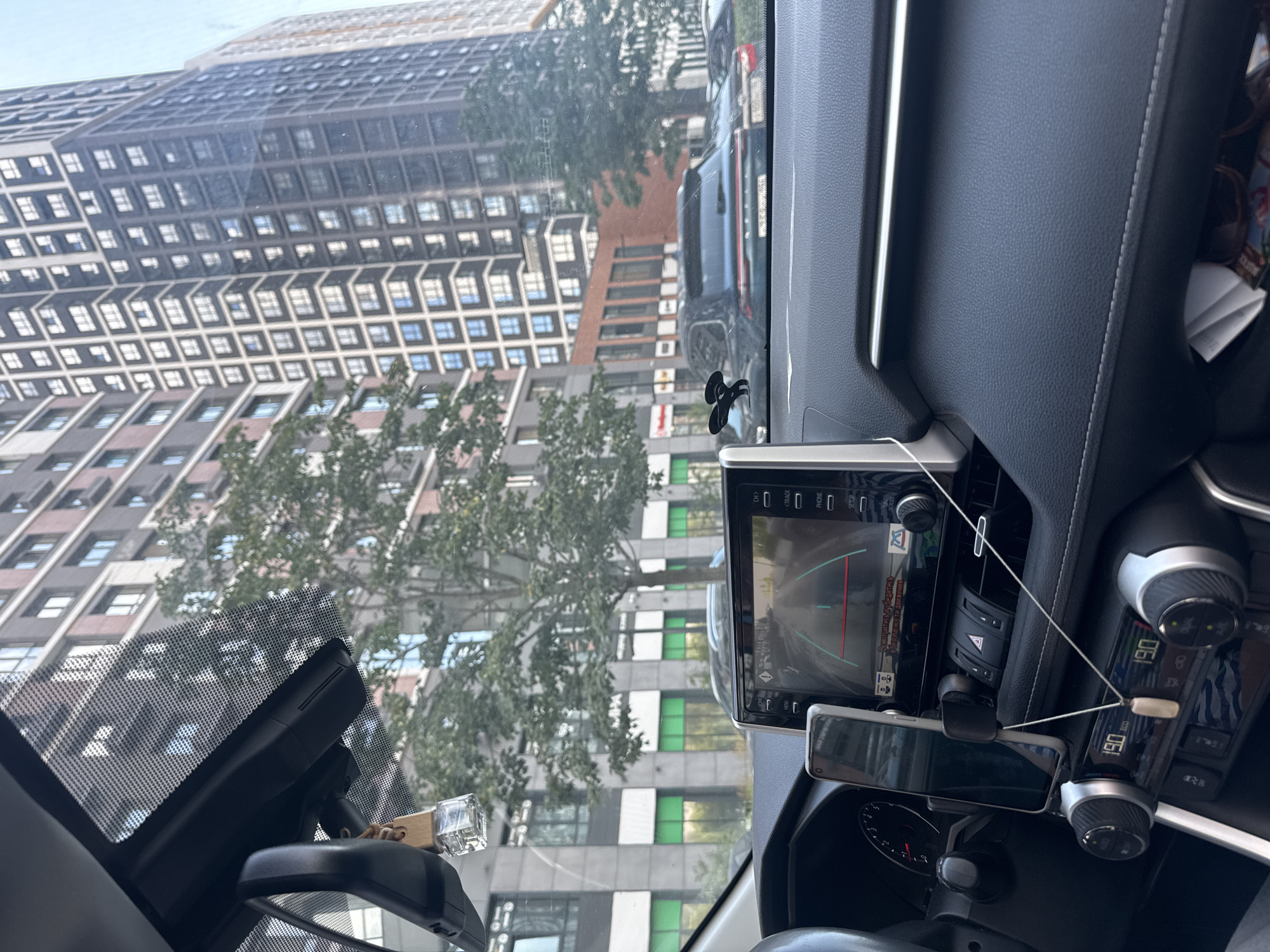The image captures the interior of a car, looking out through the windshield. The car's dashboard is visible, with a touchscreen display showing a rearview camera feed. A smartphone is mounted on the dashboard. Outside the car, a street scene unfolds with modern apartment buildings and trees. The buildings appear to be tall and contemporary, with numerous windows. Several cars are parked or driving on the street. The weather seems to be clear.