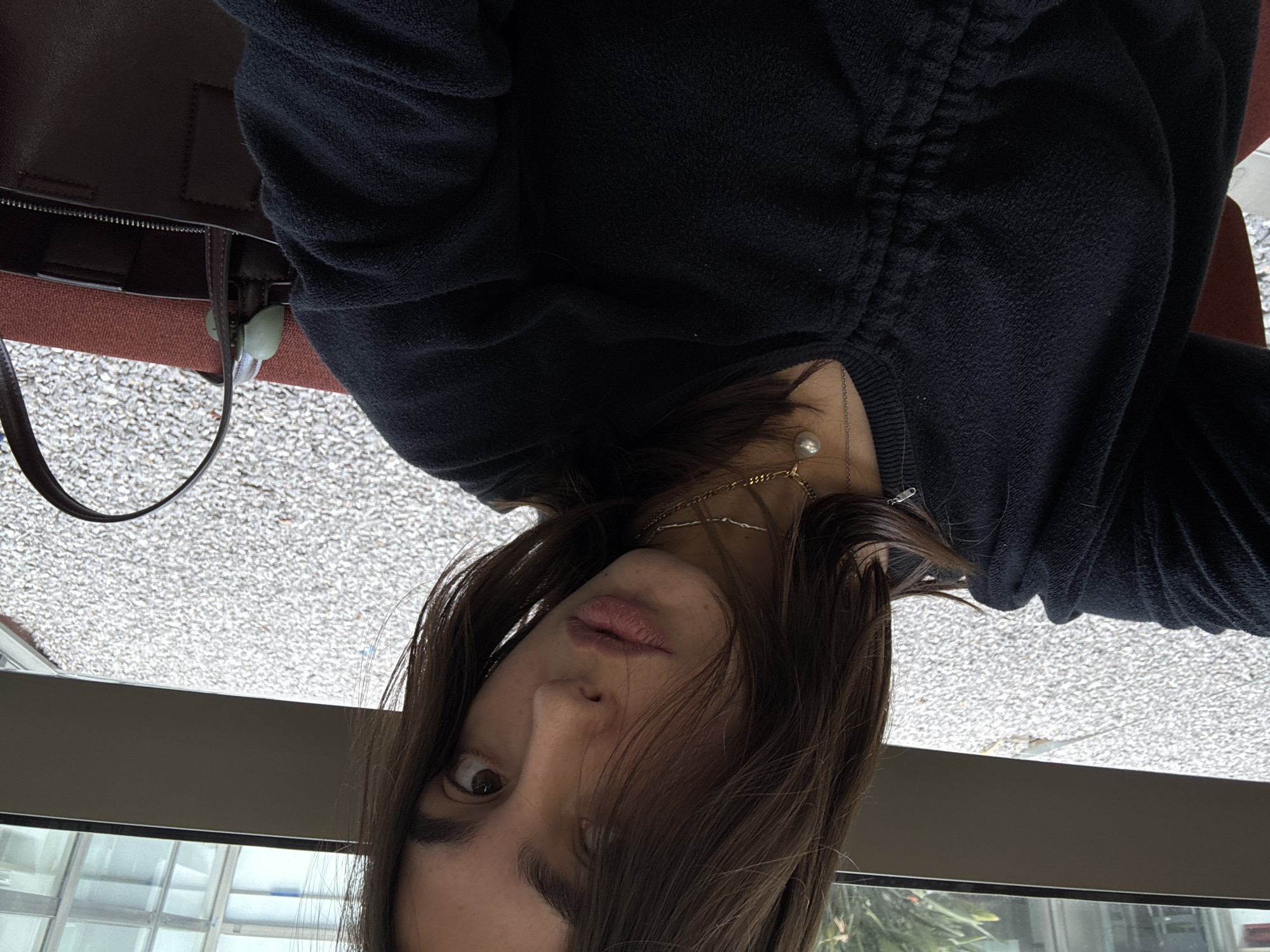 The image shows a close-up, slightly angled portrait of a young woman looking directly at the camera. She has long, dark brown hair that frames her face, and her expression is neutral with a hint of curiosity. Her lips are slightly parted.
She is wearing a black, textured, long-sleeved top, possibly a sweater or sweatshirt, with a gathered detail visible on the shoulder/sleeve. Around her neck, she wears multiple necklaces: a thicker gold chain, a finer silver-toned chain, and one featuring a prominent pearl pendant. A small silver safety pin is also visible near her left shoulder.
To her left, a dark brown leather bag with a visible zipper and strap is present. A light green or mint-colored accessory is clipped onto the bag's strap. The background suggests an outdoor or brightly lit area viewed through a window. A grey, speckled surface (possibly gravel or pavement) occupies the mid-ground, with a dark horizontal structure (like a window frame or railing) below it, revealing glimpses of greenery and architectural elements, such as glass panes. The lighting is bright, indicating it is daytime. There is no visible text in the image. The scene appears to be set in an urban or semi-urban environment, consistent with the provided context of Huixquilucan de Degollado, Mexico.