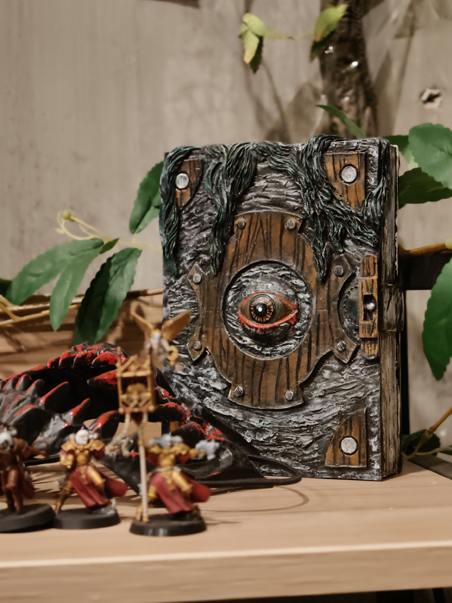 A detailed, eye-level, medium shot captures a collection of miniature figures and a decorative, occult-themed book displayed on a wooden shelf. The book, positioned in the right foreground, is rectangular and appears to be made of dark, textured material with wooden accents. Its cover features a prominent, stylized eye with a red iris and black pupil, surrounded by wooden planks and metal rivets. Dark, tangled strands resembling vines or hair drape over the top of the book.
To the left of the book are three miniature figures, painted in shades of gold, red, and brown, standing on black circular bases. They are positioned in a shallow arc, with the leftmost figure closest to the viewer. One figure holds a pole with a banner or standard. Behind these figures is a black and red object, possibly a miniature dragon or a piece of terrain, with sharp, angular protrusions.
The background consists of a neutral-colored, textured wall. Several artificial green leaves on thin stems are visible, some extending from behind the book and others from the upper left of the frame. The lighting is soft, with the primary light source seemingly coming from the front, casting subtle shadows. The overall scene suggests a display or collection of tabletop gaming miniatures and related decorative items. There is no indication of the city of Da Nang, Vietnam, in this image.