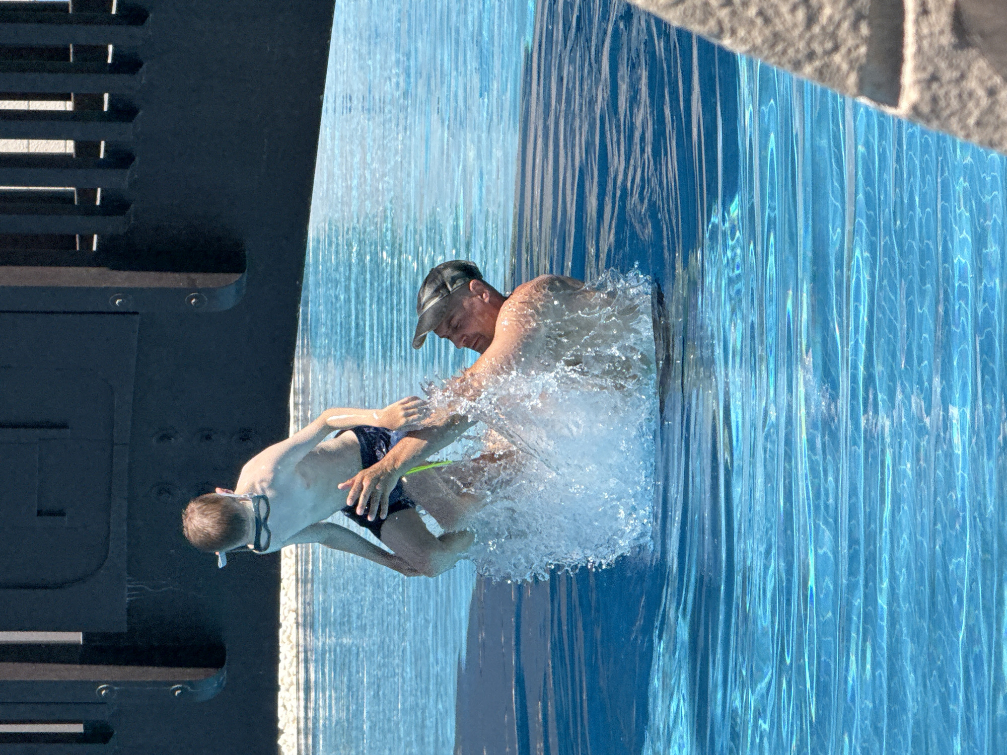 The image shows a man in a baseball cap holding a young boy in a swimming pool. The man is wearing a blue swim trunk and the boy is wearing swimming goggles and a blue swim trunk. They are both submerged in the water, with the boy being held by the man. Water is splashing around them. Behind them, there is a dark structure with horizontal slots, possibly a part of the pool's edge or a diving platform. To the right of the pool, there is a textured surface that appears to be stonework, suggesting an outdoor setting. The water is clear and blue, with sunlight reflecting off its surface, indicating it is daytime. The splashing water and the interaction between the man and boy suggest a playful moment. The context provided mentions Dubai, United Arab Emirates, which is known for its numerous swimming pools and water attractions. There is no visible text in the image.
