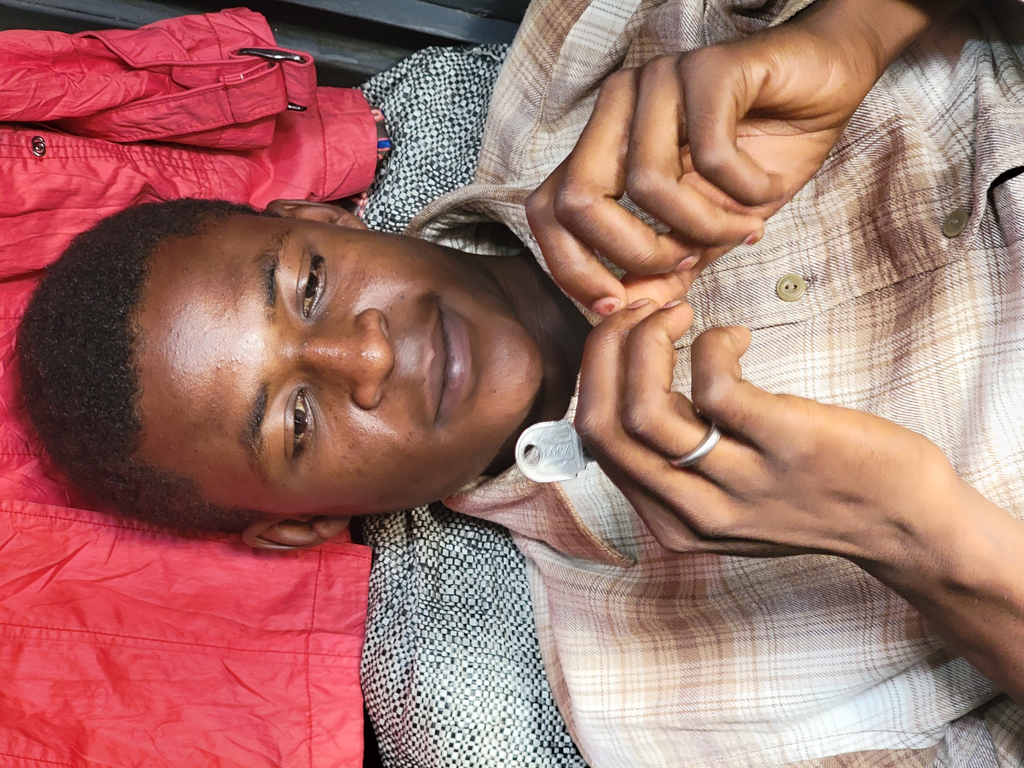 The image shows a person lying down, possibly on a bed or cushioned surface. The person has dark skin and short hair. They are wearing a light brown plaid shirt with the top buttons undone, revealing a key hanging from the shirt. A silver ring is visible on their left hand. To the left of the person, there is a red jacket. The background includes a black and white patterned fabric. The location cannot be ascertained from the image and the location information is thus irrelevant.