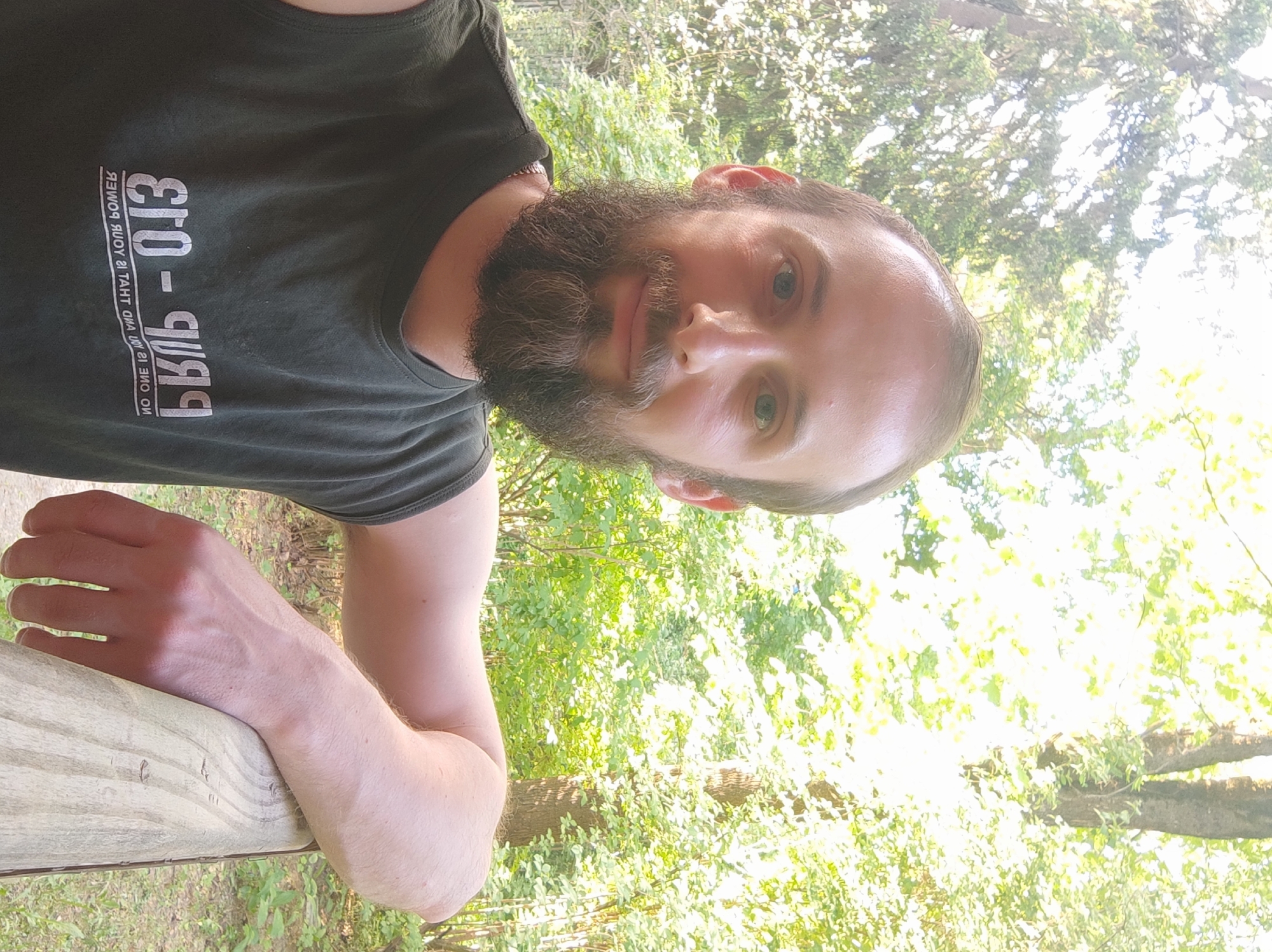A medium-angle close-up shows a man with fair skin, a dark beard, and light-colored eyes. He is wearing a dark tank top with reversed white text on the left side of the chest. The background is bright and features green foliage and tree trunks. The man is resting his arm on a light-colored wooden railing. The image does not provide enough information to identify the location as Kratovo, Russia.