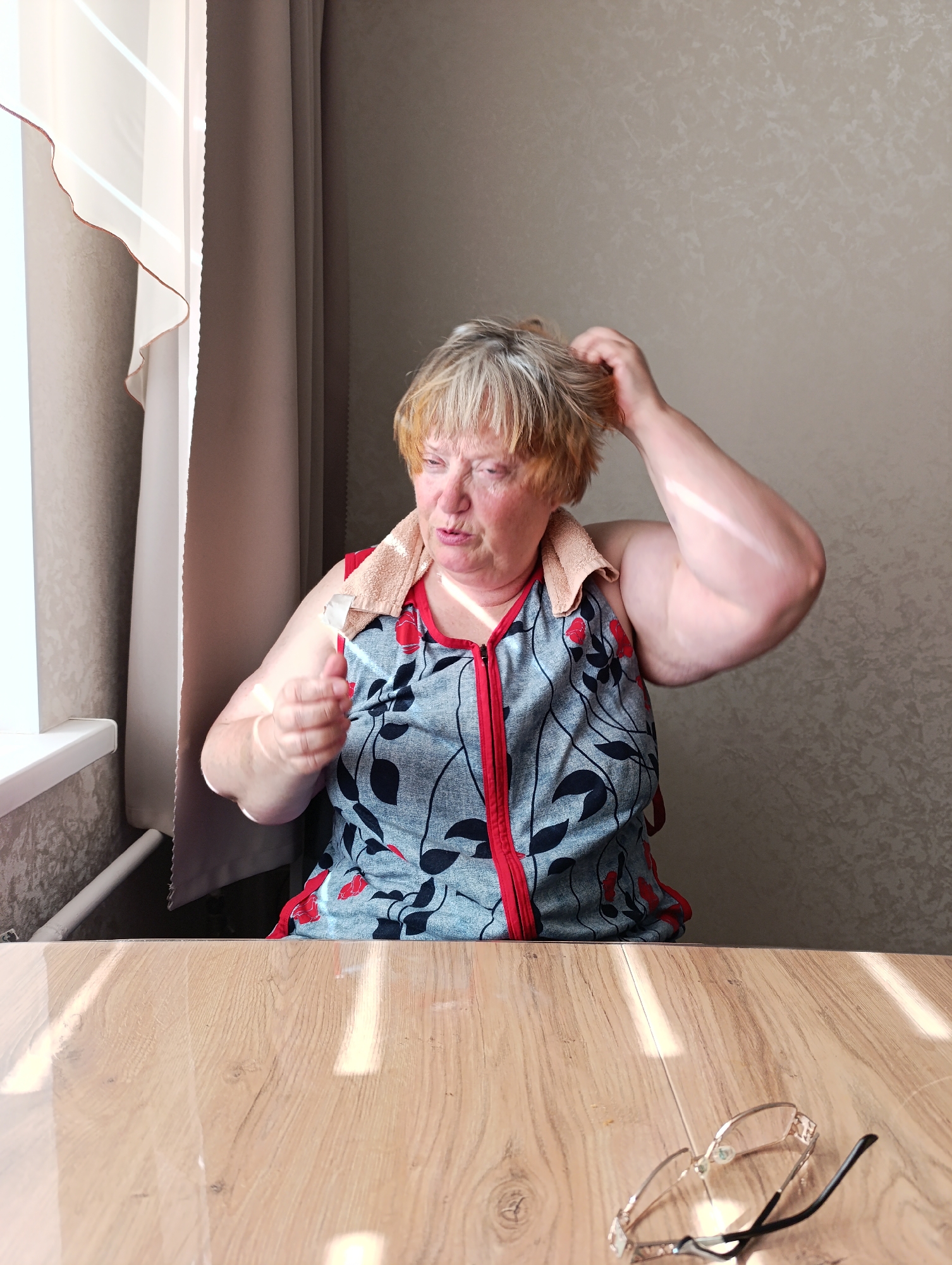 A middle-aged woman with short blonde hair, wearing a gray sleeveless top with a red zipper and a floral pattern, and a towel draped around her neck, is seated at a wooden table indoors. She is looking downwards and touching her head with her right hand, while her left hand is extended forward, holding a white object. Sunlight is casting strong diagonal shadows across the table and the woman's arm. In the background, sheer white and brown curtains are visible next to a window. On the table, in the foreground, a pair of eyeglasses with gold-colored frames and black earpieces are lying face down. The overall setting appears to be a domestic interior in Tugulym, Russia.