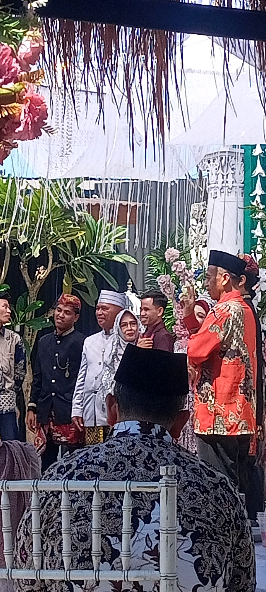 The image captures a formal gathering, likely a wedding or cultural event, outdoors in what appears to be a decorated area. Several individuals are present, all dressed in traditional attire. In the foreground, the back of a person wearing a black peci and a patterned shirt is visible, seated behind a white ornate chair.
To their right and slightly further back, a man in a vibrant red shirt with a batik pattern and a black peci is facing left, appearing to be speaking or gesturing. He is positioned next to a woman in a red hijab and a patterned dress. To the left of this couple, a man in a white traditional outfit and a striped peci stands with a slight smile, next to a woman in a patterned hijab and glasses. Further to the left, a young man in a dark jacket with a patterned sarong and a red, patterned peci is looking to his right. Another person, partially visible on the far left, is wearing a patterned shirt and a head covering.
The background features lush green foliage, floral arrangements in shades of pink and white, and decorative elements such as white fabric drapes and what looks like a white ornamental structure with intricate carvings. A thatched roof structure is visible above, with hanging decorative elements. The overall impression is of a festive and culturally significant occasion taking place in a well-decorated outdoor venue. The image is taken from a slightly low angle, looking up at the subjects.