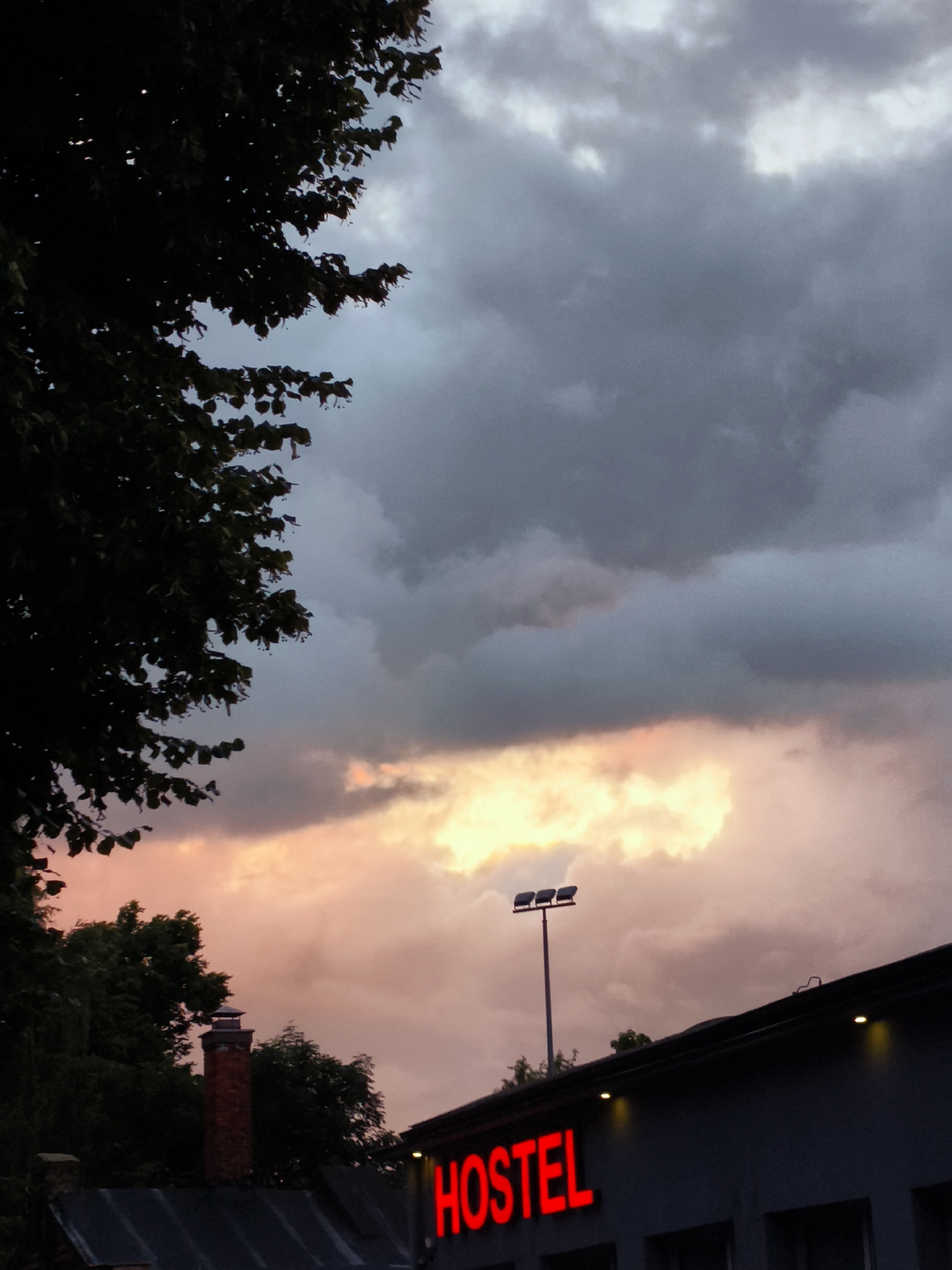 The image shows a dark, overcast sky with a patch of bright, glowing clouds illuminated by the setting sun. In the foreground, silhouetted against the sky, are the dark branches of a tree on the left. Below the clouds, a building with a dark roof is visible. A red brick chimney stands prominently on the left side of the building. The word "HOSTEL" is illuminated in bright red neon letters across the front of the building. There is also a tall pole with three floodlights on it, positioned above the hostel sign. The scene appears to be an exterior shot taken at dusk or during stormy weather. This hostel is located in the district of Wrzeszcz in the city of Gdańsk, Poland, specifically on ulica Mickiewicza.
