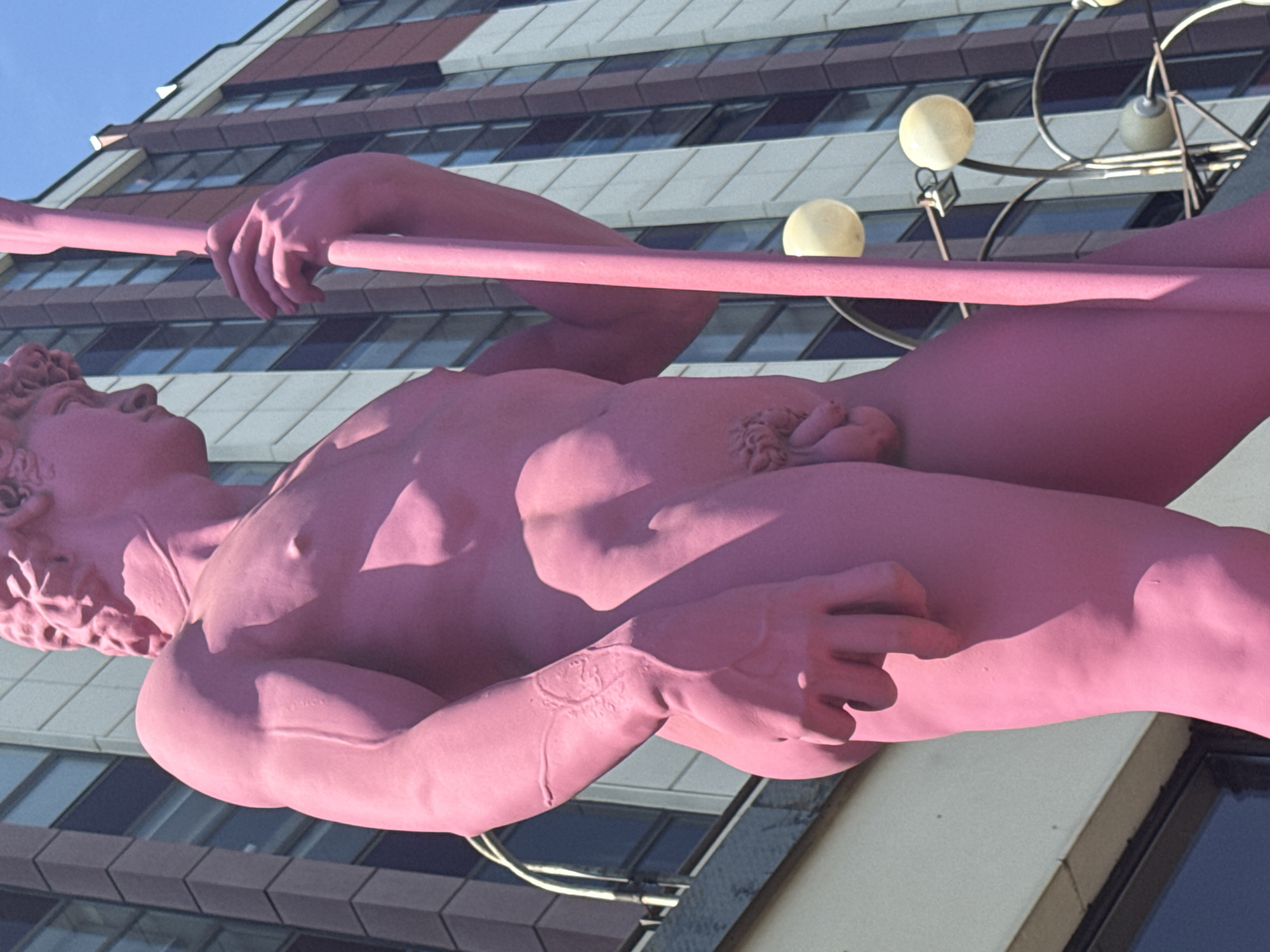 The media file shows a close-up, low-angle shot of a large, bright pink statue of a nude male figure holding a long, thin rod. The statue is made of a smooth material, and its musculature is clearly defined. The figure's head is turned to the left, with eyes looking upward. His left hand is gripping the rod, and his right arm is bent, with his hand resting on his torso.
The background is a modern building with horizontal bands of dark and light gray panels, interspersed with rows of windows. Above the building, a clear blue sky is visible. The lighting suggests it is daytime, with the sun casting shadows on the statue, highlighting its form.
In the upper right of the image, there is a decorative metal structure with several spherical, light-colored objects attached, possibly lights or ornaments. These are partially obscured by the statue. The statue is positioned in a way that it appears to be extending out from or supported by the building.
The statue is the primary subject, and its bright color and prominent placement suggest it is a public art installation. The overall impression is one of grandeur and perhaps a surreal, playful aesthetic due to the unusual color of the statue. No text is visible in the image.