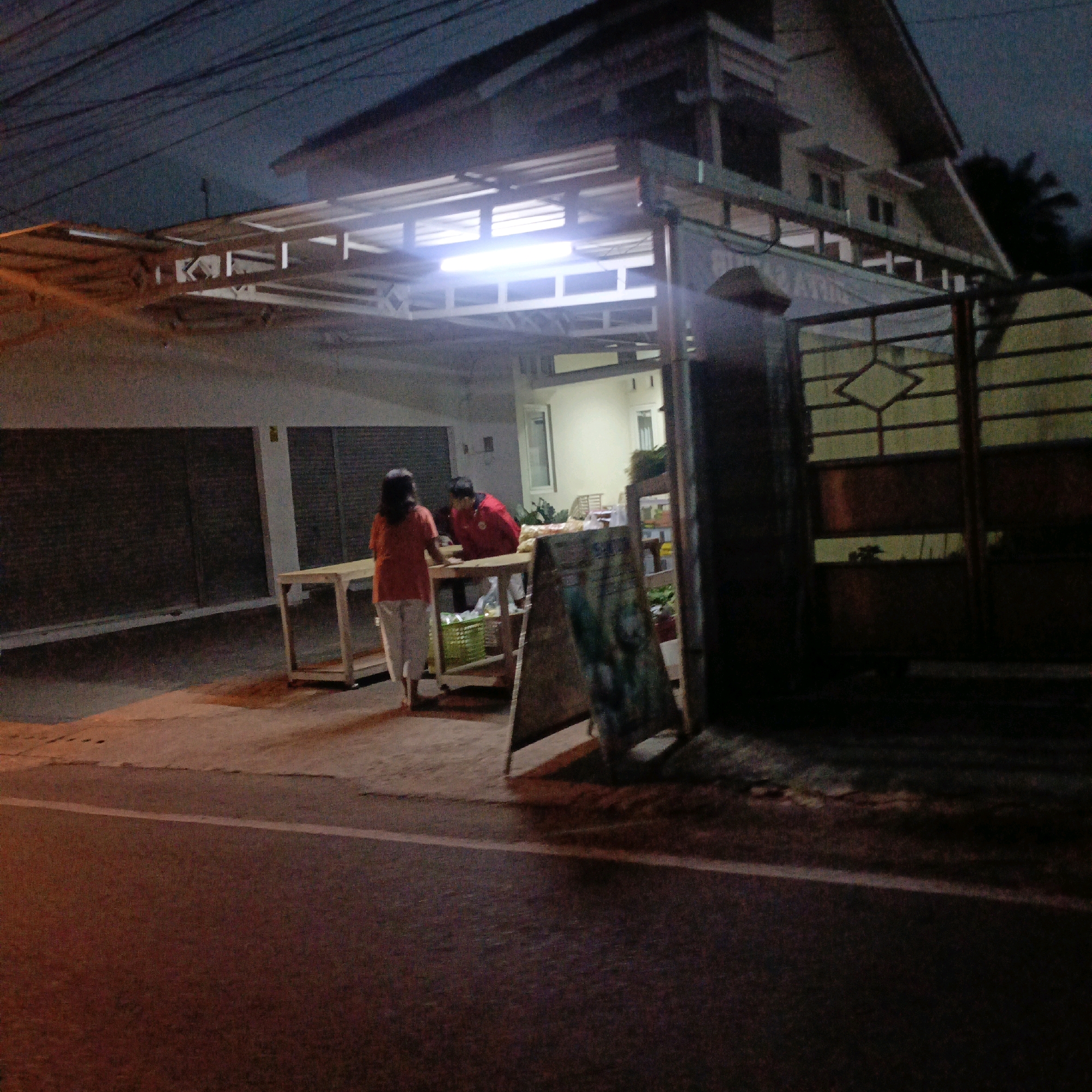 The image depicts an outdoor scene at dusk, likely in Salatiga, Indonesia, showing a street-side stall or small shop. Two individuals are present: a woman in an orange shirt and light-colored pants stands to the left, and a man in a red shirt is on the right, leaning over a wooden table. The table is laden with goods, possibly food items or produce, and a green basket is visible underneath. An A-frame sign with illegible writing stands in front of the table. In the background, a building with a sloped roof and illuminated windows is visible, along with what appears to be a large metal gate. The lighting is dim, suggesting it is either early morning or late evening, with artificial lights from the stall and building illuminating the scene. The street in the foreground is paved with a white line marking.