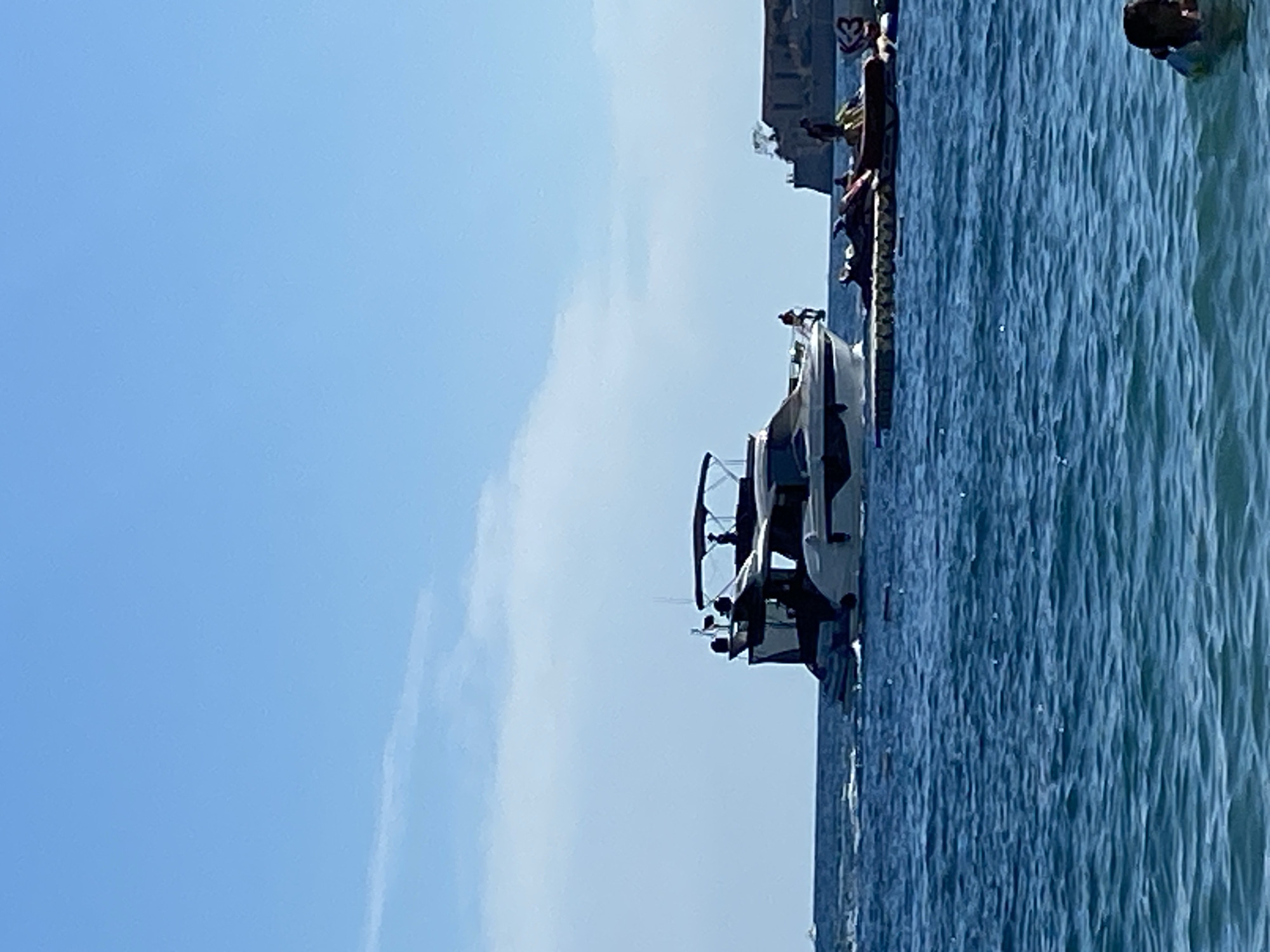 An eye-level, medium shot captures a bright, sunny day at a beach in Budva, Montenegro. The foreground is dominated by deep blue, rippling ocean water, with a person's head and shoulders visible in the lower right corner, partially submerged. In the mid-ground, a sleek white and black motor yacht is moving across the water, with several people visible on its deck. To the right of the yacht, a floating platform is laden with several jet skis and more people. In the background, a historic stone fortress with multiple buildings and turrets sits on a rocky outcrop overlooking the sea. The sky above is a clear, light blue with faint white clouds.