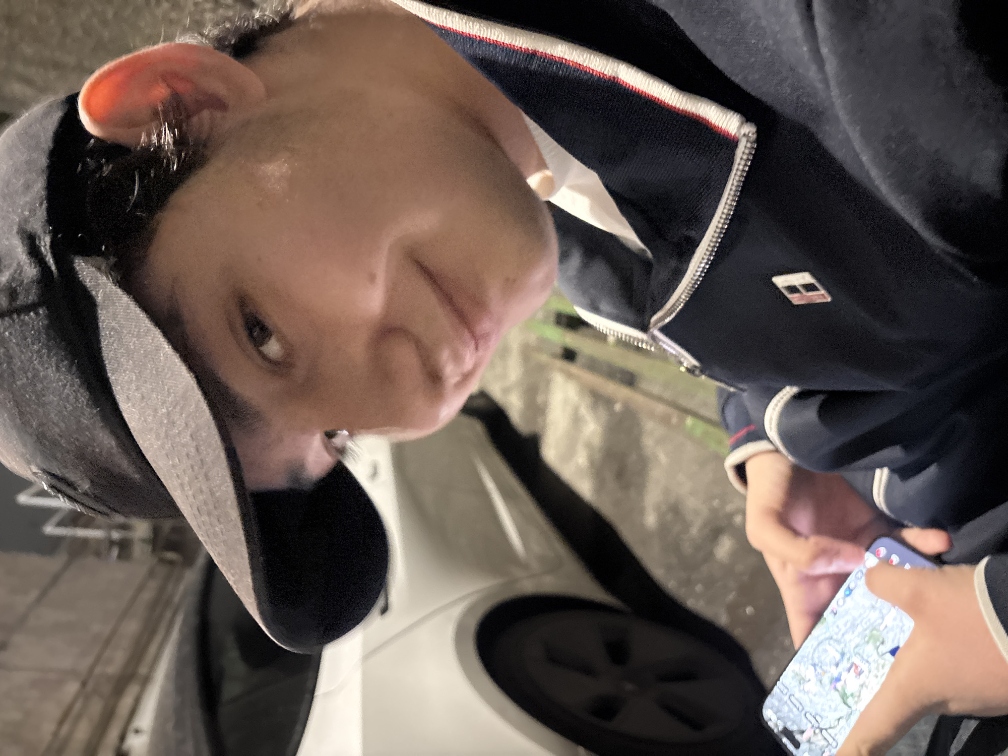 A person wearing a dark jacket with white and red trim and a black baseball cap is holding a phone in their hands. The phone screen displays a map-like interface with various icons. The person is positioned near a white vehicle, possibly a car. The location appears to be outdoors, possibly at night, judging by the lighting. I cannot determine the specific location in Yerevan, Armenia, without additional context or details from the image.