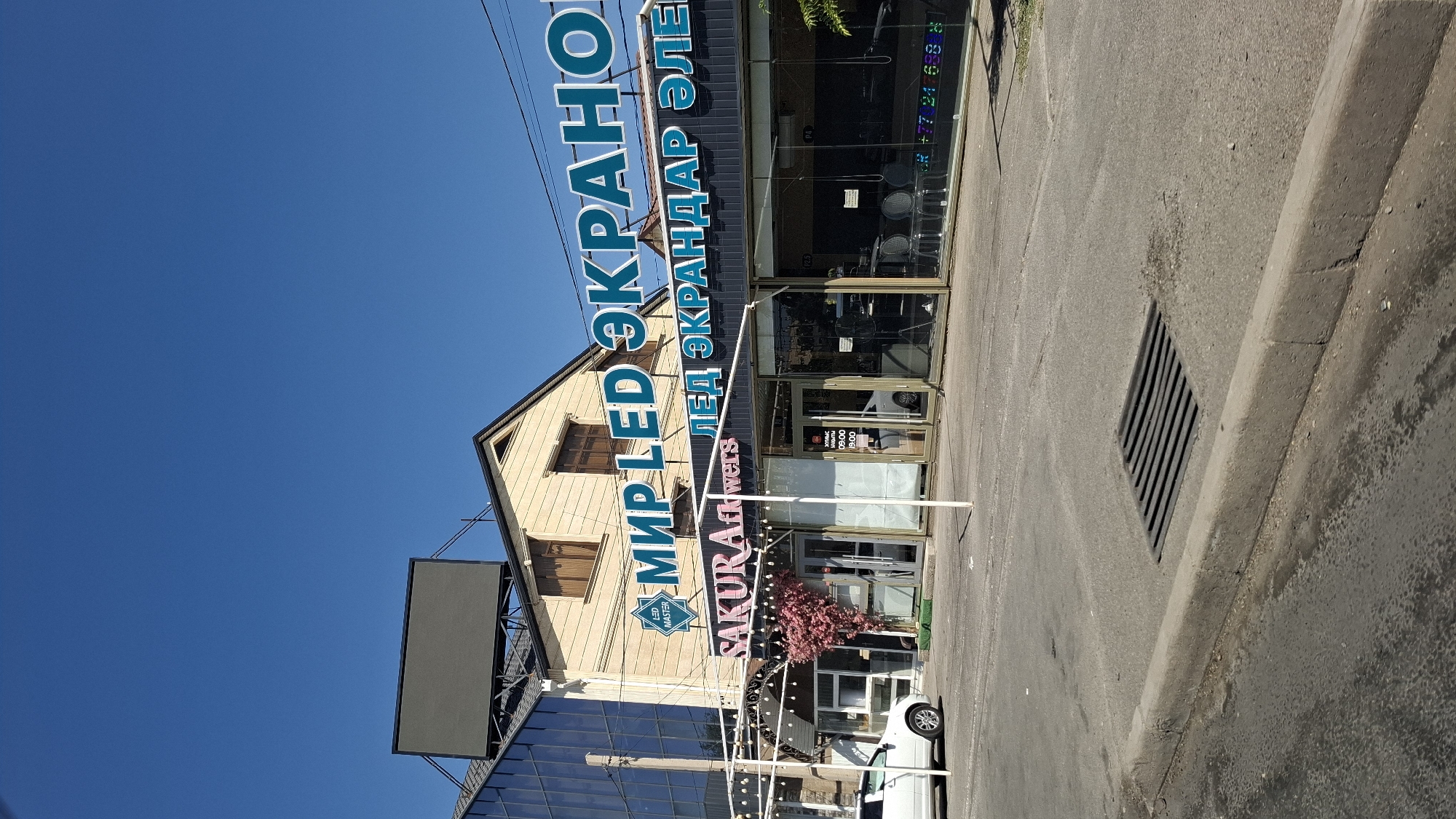 The image shows a street-level view of a commercial building in Shymkent, Kazakhstan. A large, dark billboard is mounted on the roof of the building. The building has a glass facade on the left and a more traditional structure with windows on the right. A prominent sign in blue letters reads "МИР LED ЭКРАНО" (World LED Screens). Below this, another sign in blue letters says "ЛЕД ЭКРАНДАР ЭЛЕ" (LED Screens Ele). A smaller pink sign reads "SAKURA flowers". A white car is parked on the left side of the street in front of the building. The street is paved, and a storm drain is visible in the foreground. The sky is clear and blue. The overall impression is one of a retail or service establishment in a well-lit urban environment.