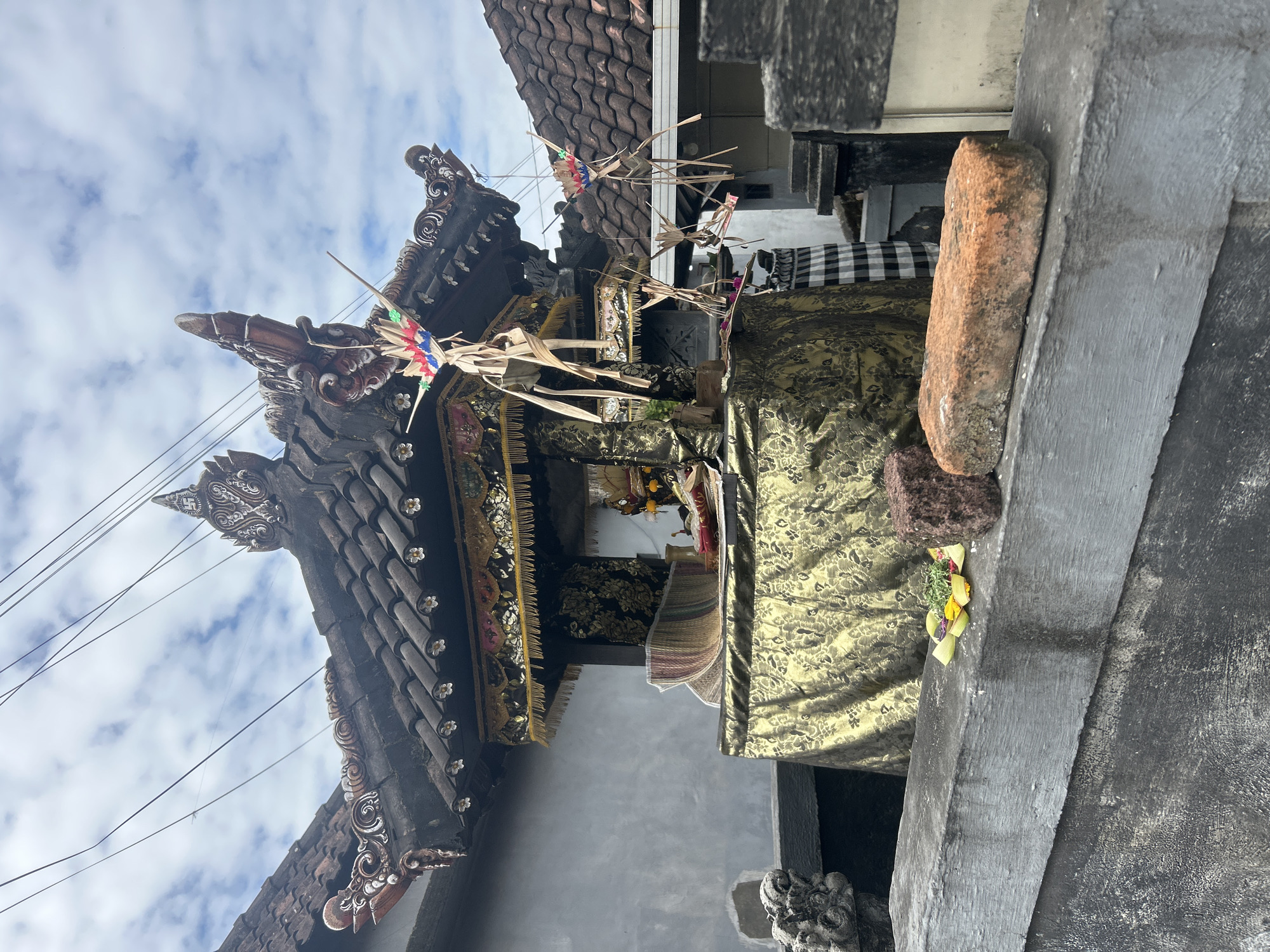 The image depicts an outdoor altar, likely a household shrine, in Denpasar, Indonesia. The altar is adorned with gold and black patterned fabric, with a woven mat and offerings of food and flowers placed on its surface. The structure is topped with a tiered, dark-tiled roof featuring ornate carvings and what appear to be small decorative flowers. Several palm frond decorations hang from the roofline. In the foreground, a concrete wall runs horizontally, with two objects resting on it: a reddish-brown brick and a dark, rough-textured stone. Behind the altar, a light-colored wall is visible, and the roof of an adjacent building with red tiles can be seen. The sky is partly cloudy. The presence of the altar and the architectural style suggest a Balinese Hindu context.