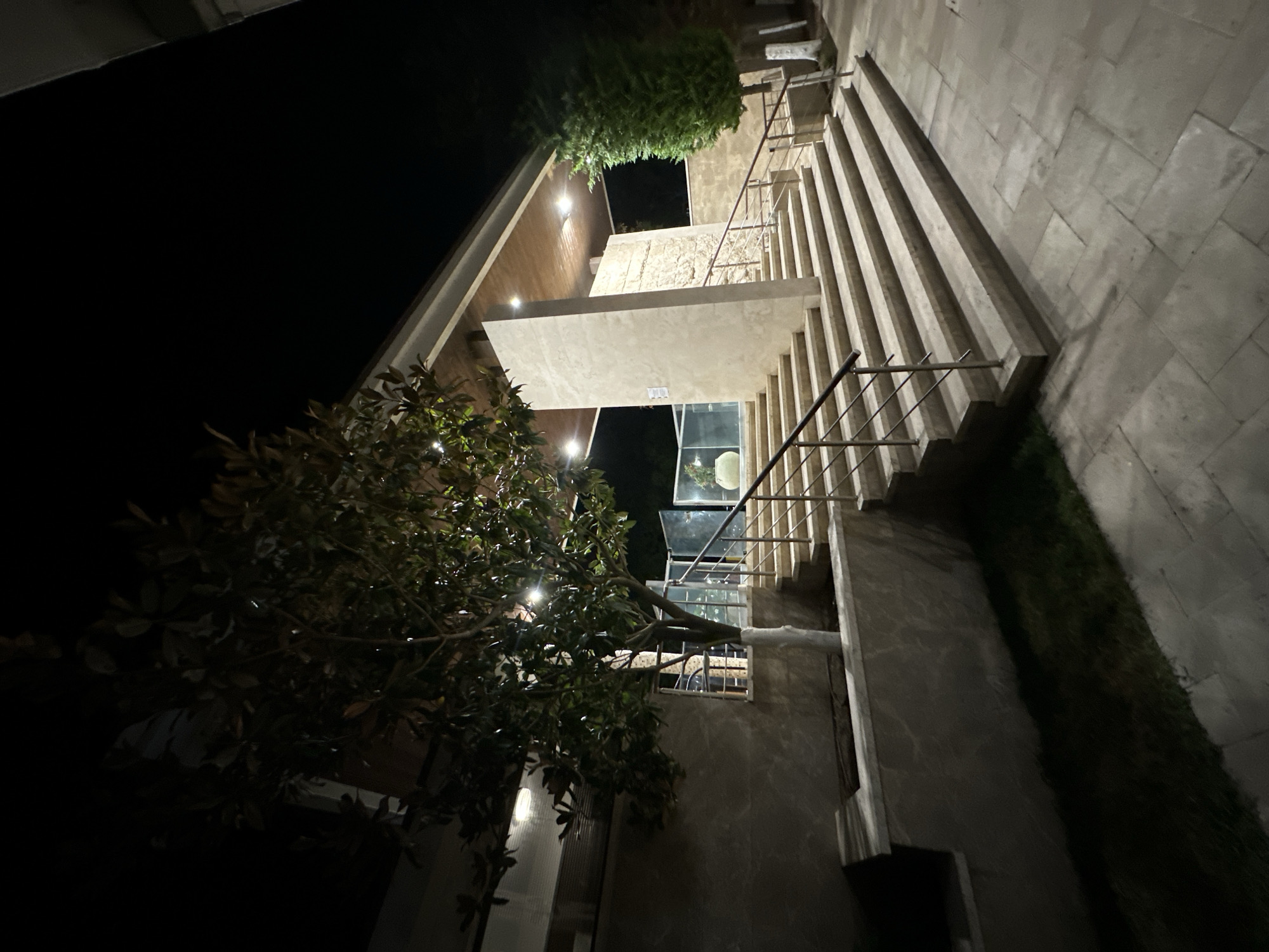 The image shows an outdoor stairway leading up to a building entrance at night. The stairway is made of light-colored stone or concrete with metal railings on both sides. The entrance is covered by a canopy with wooden panels on the ceiling and recessed lighting. A large tree with green leaves and some decorative string lights is positioned to the left of the stairs. To the right of the stairs, there is a tall, narrow green tree. The area is paved with large stone tiles. The background is dark, indicating nighttime. The architecture suggests a modern, possibly residential or commercial building. The scene is calm and devoid of any people. The location is identified as being in Sozopol, Bulgaria, suggesting a context of a specific place within that city.