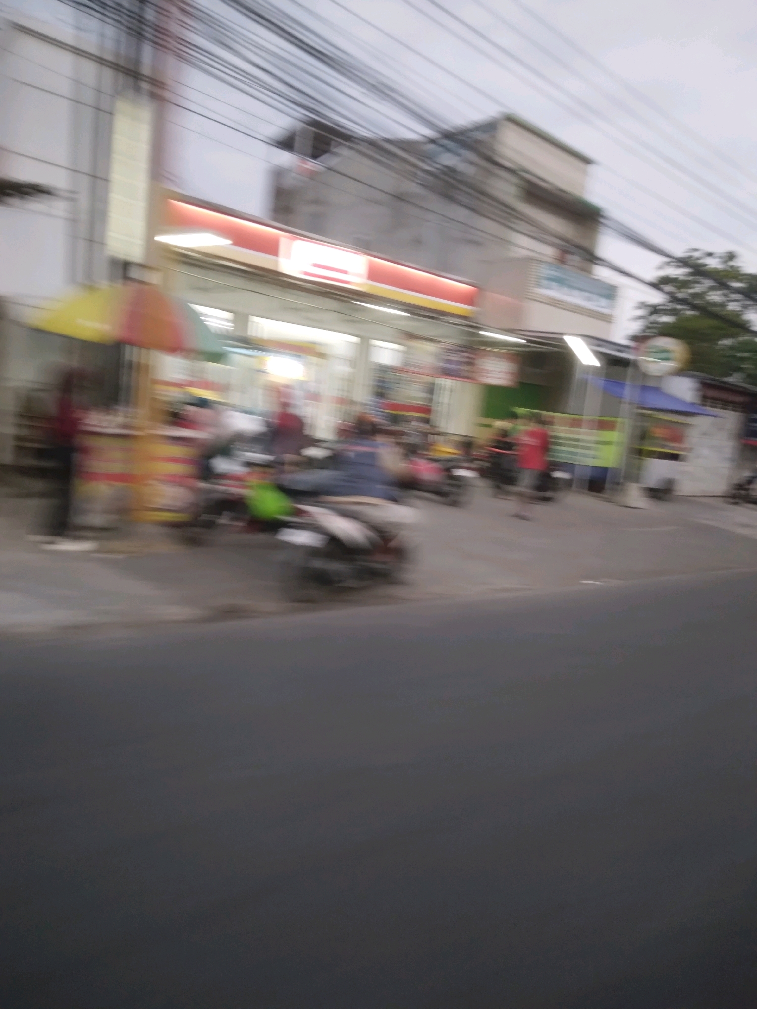 The image depicts a street scene in Sokaraja, Indonesia, likely captured from a moving vehicle due to motion blur. In the background, a brightly lit storefront, possibly a convenience store or minimarket, is visible with signage that is too blurry to read. Several individuals, their forms blurred, are present around the store. Some are standing, while others appear to be near or on motorcycles, suggesting a public area with pedestrian and vehicle traffic. There is also a colorful umbrella or canopy visible on the left side of the frame, possibly part of a street vendor's setup. Power lines are prominent in the upper portion of the image, indicative of an urban or semi-urban environment. The overall lighting suggests it could be dusk or overcast weather. The foreground is dominated by a blurred asphalt road.