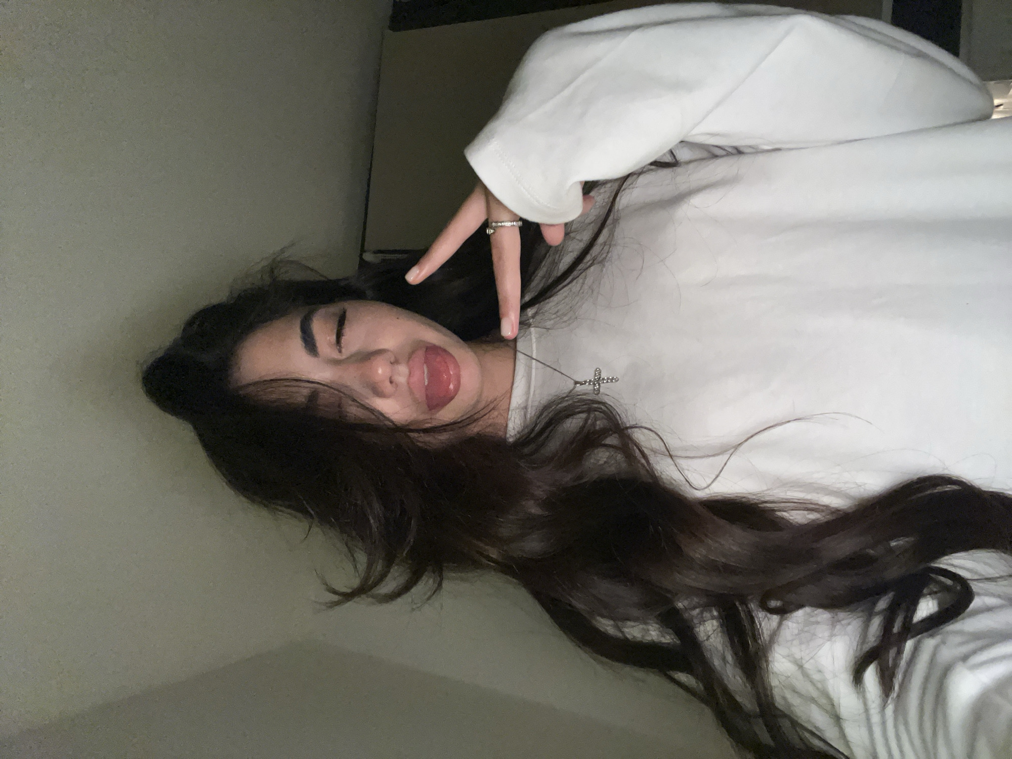 A young woman with long, dark, wavy hair is shown from the chest up, taking a selfie in what appears to be a dimly lit room. Her left eye is closed, and her tongue is sticking out. Her right hand is raised, with two fingers extended in a peace sign, and she is wearing two rings on that hand. She is wearing a white long-sleeved shirt and a silver cross pendant necklace. The background is a textured, dimly lit wall. There are no discernible landmarks or objects that would indicate the specific location within Chotěšov, Czechia.