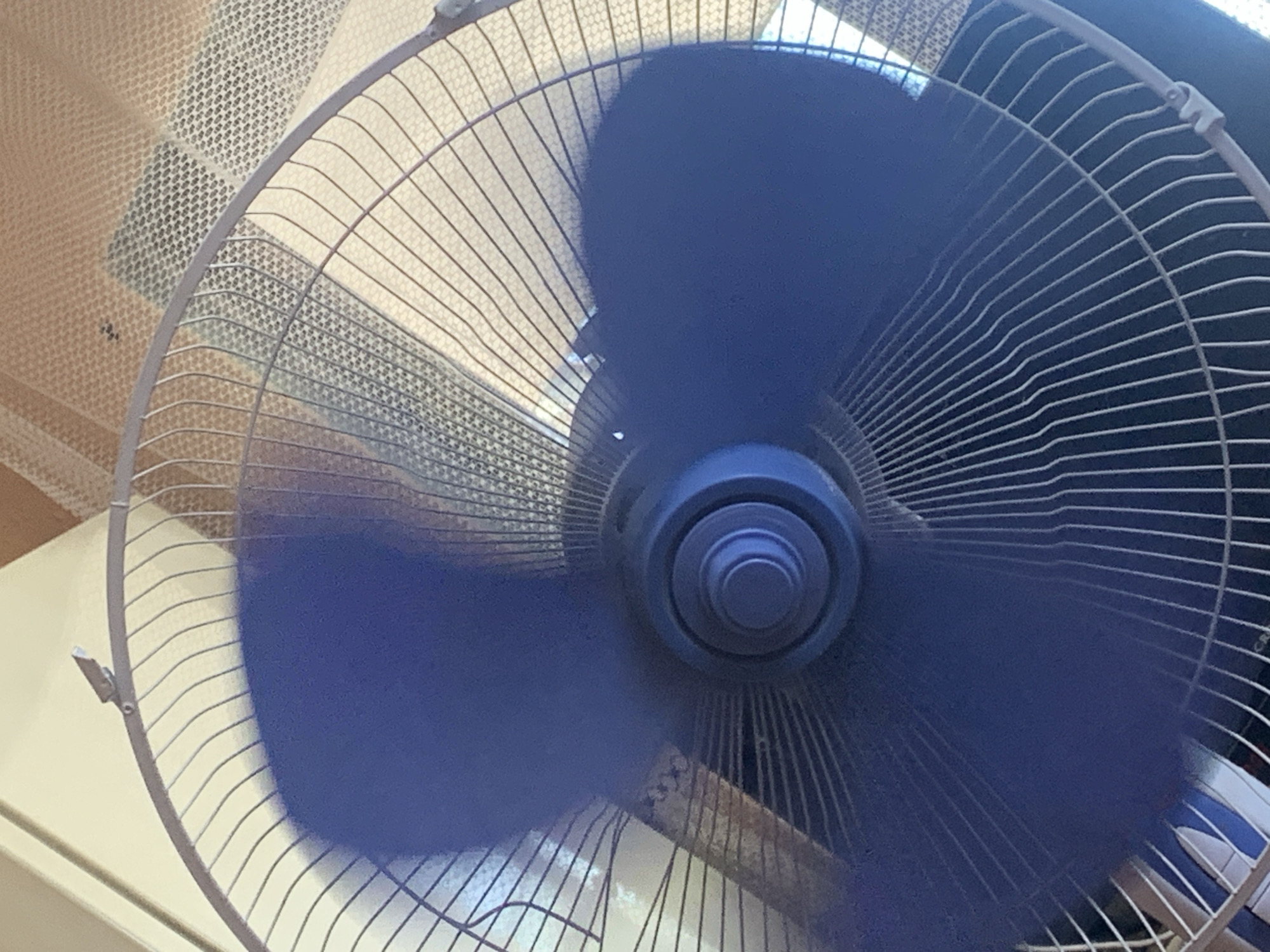 A close-up, overhead shot shows a dark blue fan with its blades blurred from rotation. The fan is positioned in front of a textured, light-colored wall or screen. The metal grating of the fan is visible, with thin, parallel wires forming a protective cage. The central hub of the fan is also dark blue, with concentric circles. The lighting appears to be natural, casting shadows on the fan blades. The image was taken in Mechkarevo, Bulgaria.