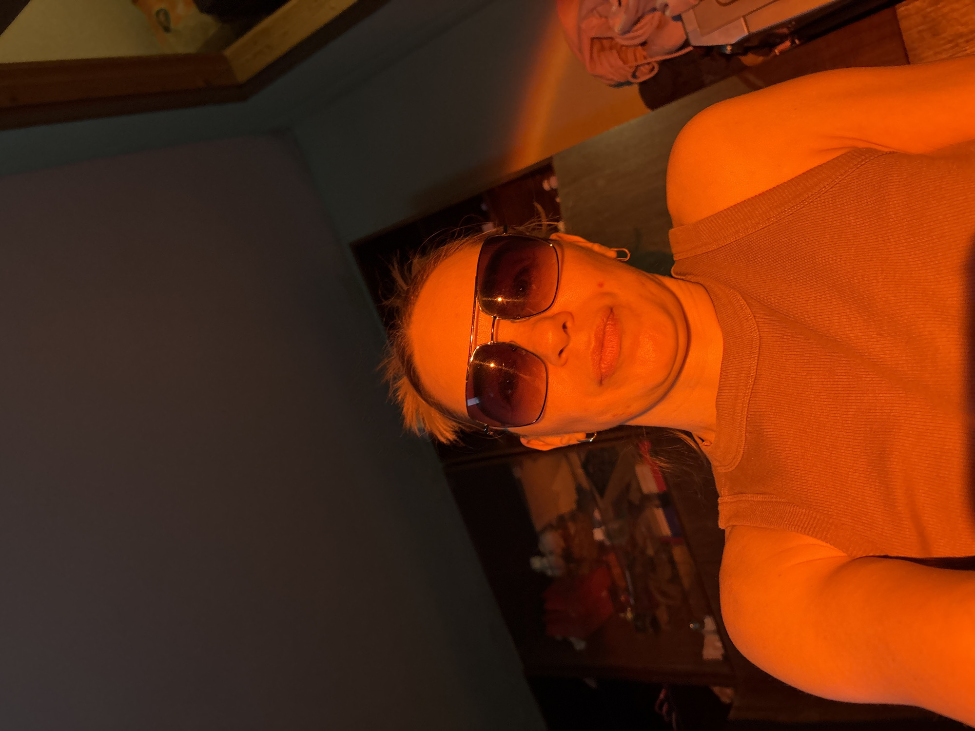 A woman with blonde hair is wearing sunglasses and a brown tank top. She is indoors. The location is Unknown, Russia. The lighting is orange.