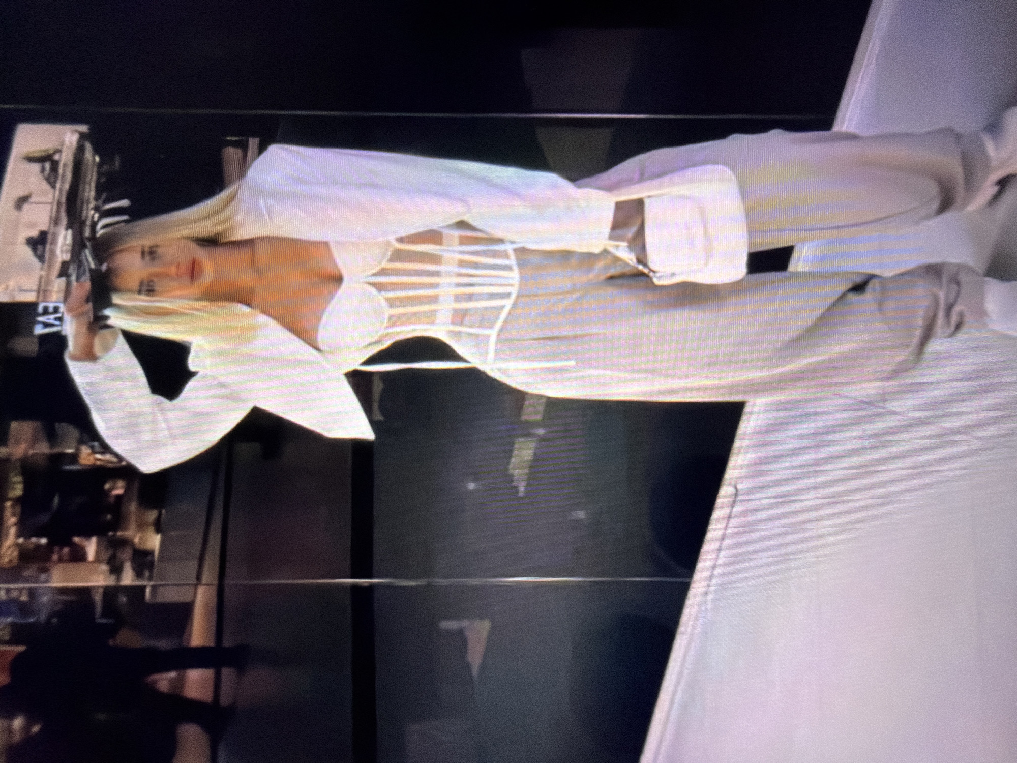 The image shows a young woman with blonde hair wearing a white corset, a white blazer, and light-colored baggy pants. She is holding her hand on her head and looking at the camera. She is also carrying a small white handbag. The background appears to be a modern interior, possibly a store, with reflections in the glass walls. There are blurry figures in the background, and what looks like store displays. The scene is lit with artificial lighting. The location is Beșalma, Moldova.