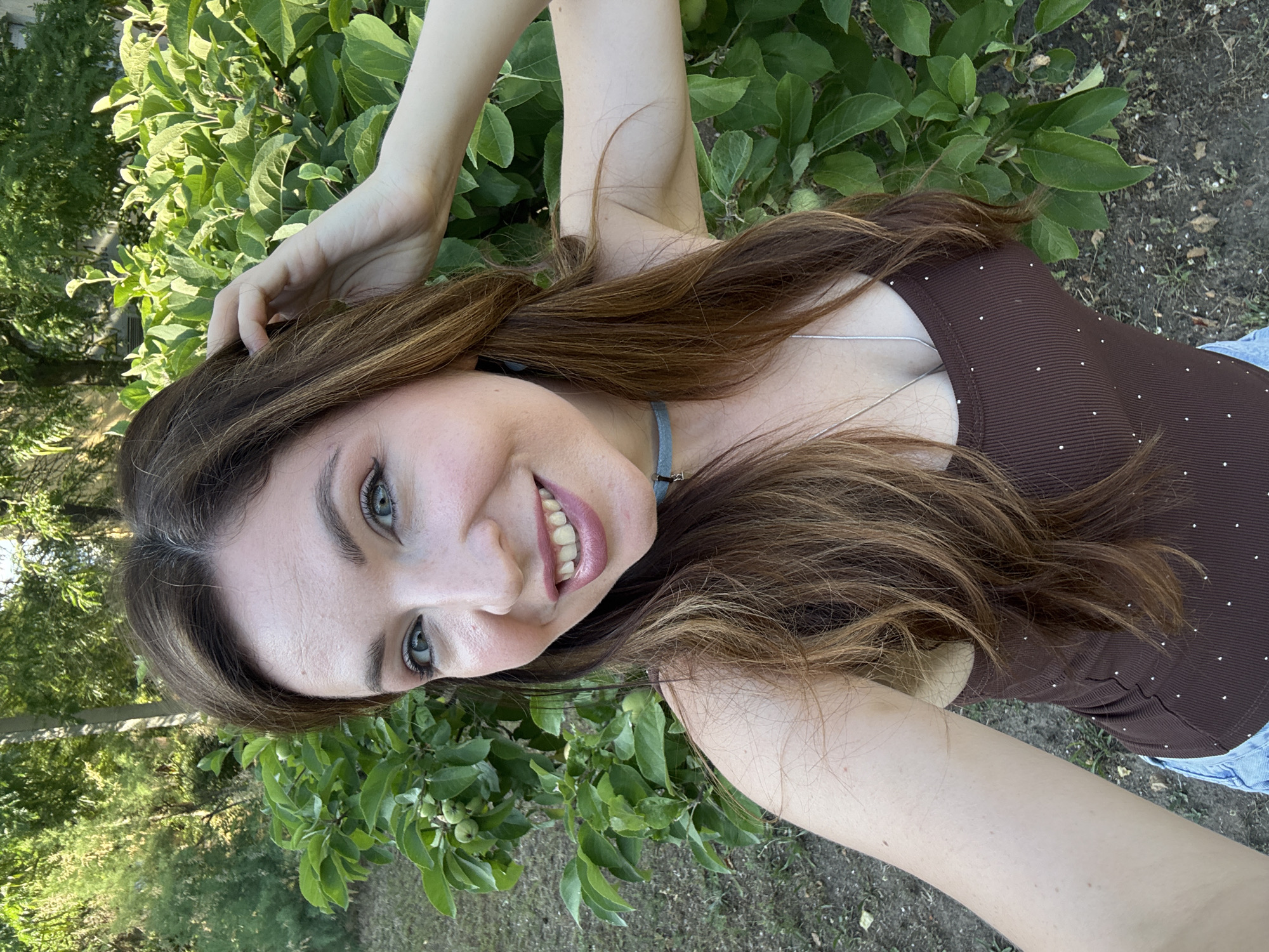 The image shows a young woman taking a selfie outdoors. She has long, brown hair and is smiling, with her head tilted to the side and one hand raised to her hair. She is wearing a brown, ribbed tank top with small white polka dots and a silver necklace. The background consists of lush green foliage, suggesting a natural outdoor setting. The lighting indicates it is daytime, possibly late afternoon due to the warm tone of the light. The ground beneath the foliage appears to be soil with some fallen leaves. The overall mood is relaxed and cheerful. There is no visible text in the image.