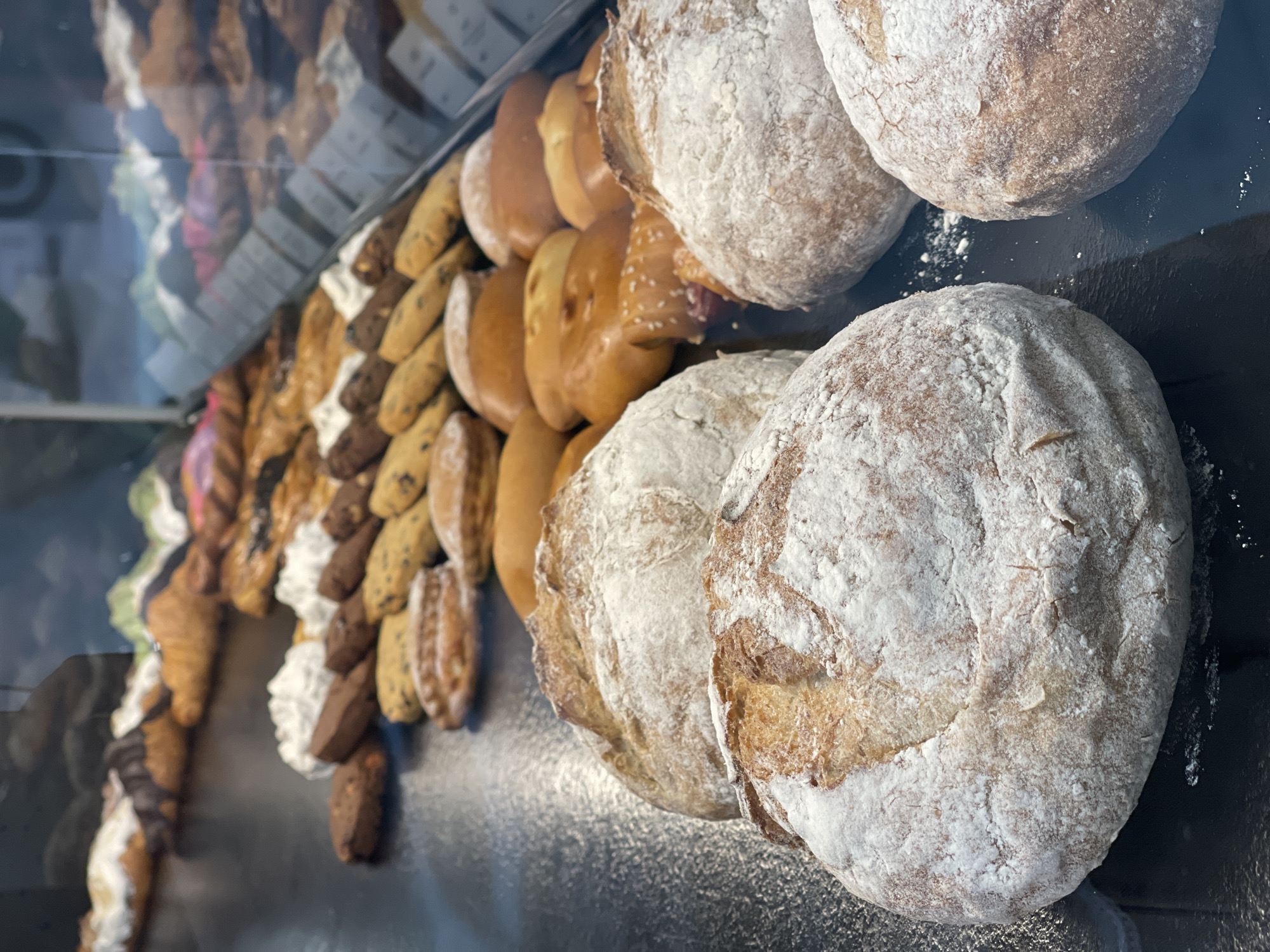 A display of baked goods is presented in a bakery. The foreground features several rustic loaves of bread, dusted with flour. Behind them, rows of cookies and pastries, including croissants and what appear to be danishes, are arranged on shelves. The overall scene is well-lit and suggests a commercial bakery setting.