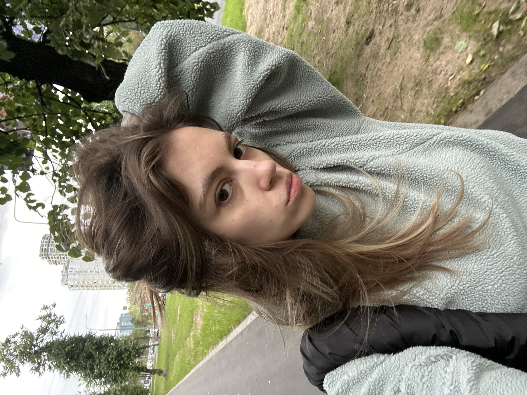This media file is a selfie of a young woman taken outdoors. She is wearing a light blue fuzzy hoodie and a black jacket. Her long, light brown hair is spread out, and she is looking at the camera with a neutral expression. Her right arm is bent, with her hand behind her head, a pose that suggests a casual and relaxed demeanor.
The background shows a paved walkway leading to a grassy area with some trees. In the distance, there are buildings, suggesting an urban environment. The buildings are modern, with some having colorful facades. The sky appears overcast, indicating a cloudy day. There are power lines visible between the trees and buildings.
The overall scene is one of a person taking a selfie in a park or a green space within a city. The time of day is not explicitly clear but appears to be daytime due to the lighting. The weather seems cool, given the clothing. No text is visible in the image. The location context suggests Moscow, Russia, which aligns with the presence of multi-story buildings and greenery in an urban setting.