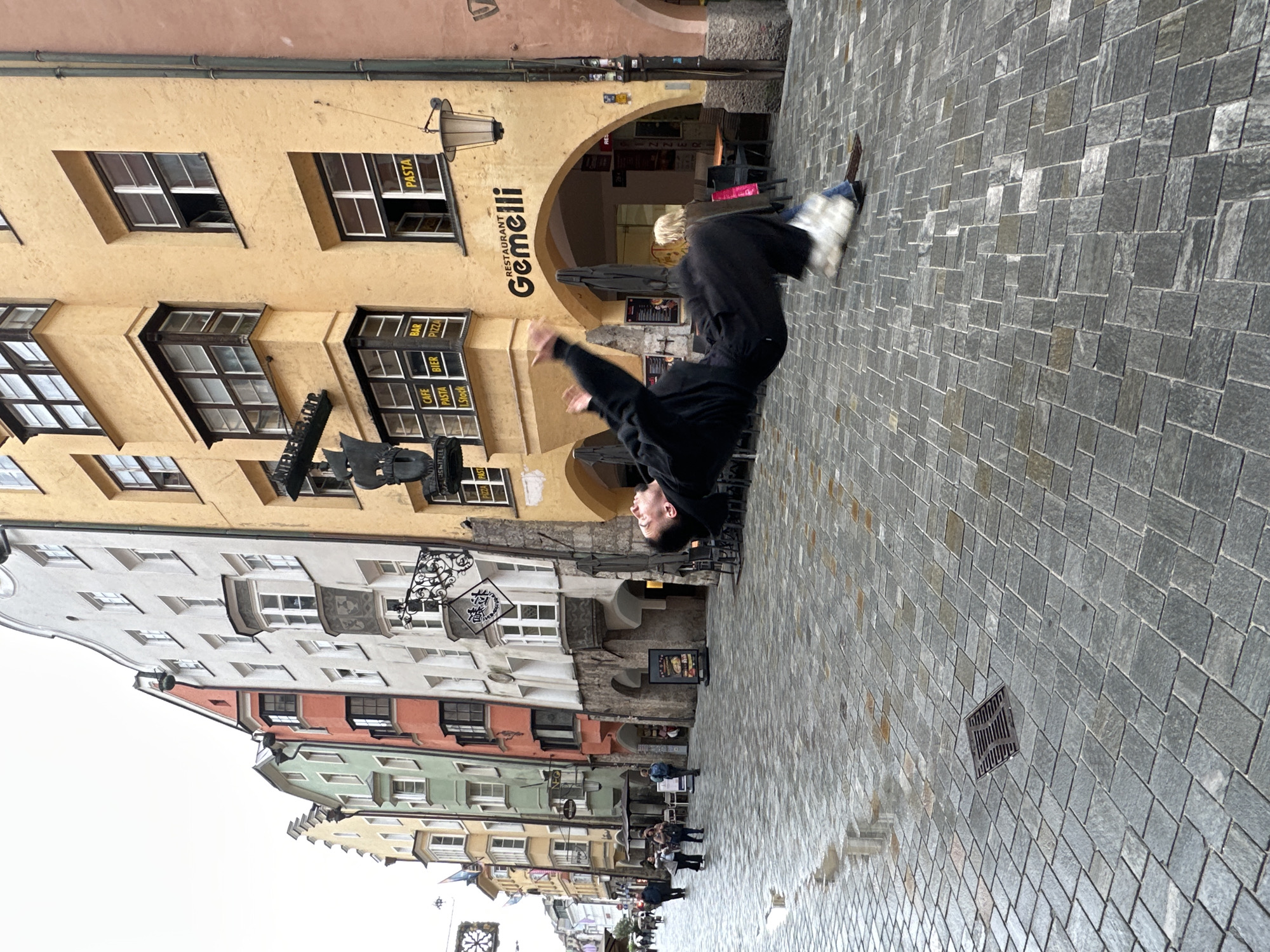 This image depicts a street scene in Innsbruck, Austria, likely on a cloudy or overcast day, as evidenced by the diffuse lighting and wet cobblestone pavement. The architecture suggests a historic European city center, with colorful, narrow buildings lining the street. The building on the right is painted a striking yellow and features arched doorways, likely leading to shops or restaurants. A sign above one of the arches reads "RESTAURANT Gemelli". Several windows are visible, some with awnings, and one window displays a yellow sign with the word "PASTA".
The focal point of the image is a person captured mid-air, performing a parkour-like acrobatic maneuver, possibly a flip or a dive. The individual is wearing dark clothing, including a hooded jacket and pants, and appears to be wearing light-colored footwear. Their body is contorted as they move through the air.
In the background, other people are visible, some walking along the street, and one person with blonde hair is seen near the yellow building. The street is paved with cobblestones, which are glistening, indicating recent rain or dampness. A metal grate is visible in the foreground, partially submerged in the wet pavement. The overall atmosphere is one of a bustling urban environment with an unexpected display of athletic prowess.