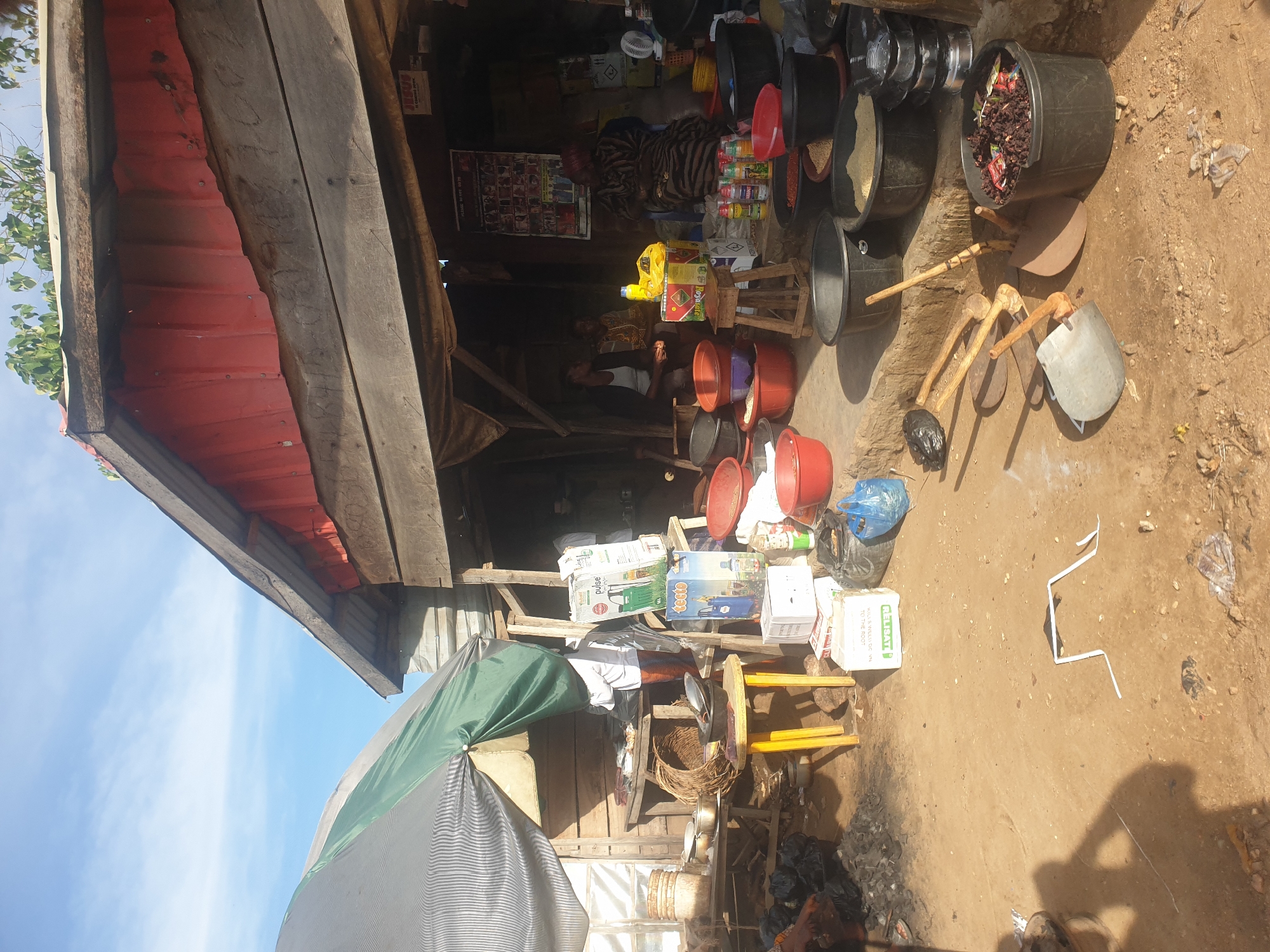 A roadside market stall in an unidentified location shows a variety of goods and tools arranged around the structure. The stall's construction includes a red-painted corrugated metal roof and wooden supports. A large, striped umbrella provides additional shade.
Inside the stall, a vendor sits amongst an assortment of merchandise, including stacks of containers, boxed goods, and buckets filled with grains and other items. The foreground displays more containers and tools such as hoes and shovels placed on the ground. The ground itself is dirt.