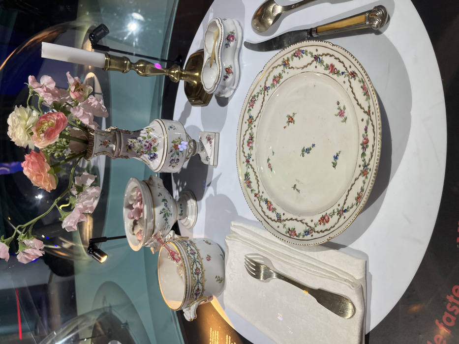 This image presents a meticulously arranged antique table setting, displayed within an exhibition at the Cité Internationale de la Gastronomie et du Vin in Dijon, France. The collection of porcelain and silverware rests on a white, semi-reflective circular surface, encased behind a clear, curved barrier.
In the foreground, a white porcelain plate, adorned with a delicate floral garland border and scattered small floral motifs in its center, is positioned centrally. To its left, a silver-toned fork with a broad handle rests upon a neatly folded white napkin. To the plate's right are an ornate knife with a light, possibly bone or ivory, handle and a silver-toned spoon.
Behind the plate, from left to right, are several additional items. A white porcelain teacup, decorated with floral patterns and a patterned rim, sits to the left. Adjacent to it is a footed white porcelain bowl with a lid, containing numerous small, pale pink and white candies. Central to the rear of the display is a tall, slender white porcelain vase, richly decorated with colorful floral patterns, holding an arrangement of artificial flowers, including pink sweet peas and orange ranunculus. To the right of the vase, a brass candlestick supports a white taper candle. The furthest item to the right is a small, two-compartment white porcelain dish, likely a salt cellar, containing a miniature golden spoon in each compartment.
The background is dimly lit with hints of blue, and reflections are visible on the clear barrier. Small, focused spotlights illuminate the individual display items. Partially visible text, including "e faste," is present on a dark surface in the lower left portion of the frame, indicating an exhibit label.