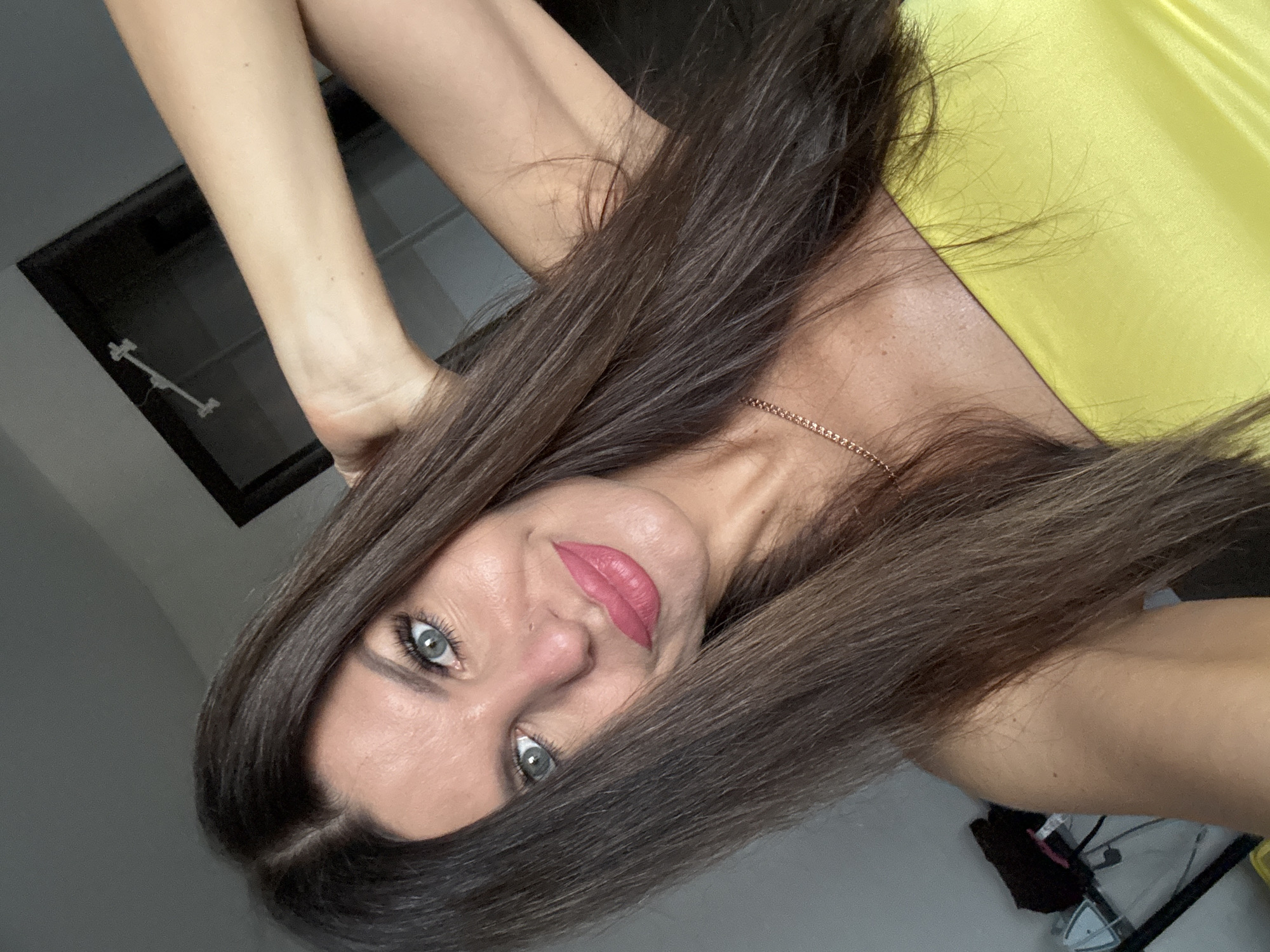 A medium shot shows a woman with long, dark brown hair, fair skin, and green eyes. She is wearing a yellow strapless top and a delicate rose gold necklace. Her right arm is raised and her hand is behind her head. The woman is looking towards the camera with a slight smile. The background is indistinct but appears to be an indoor setting with a closet and a mirror visible. The image is a selfie, suggesting the woman is taking the photo herself. The location is identified as Chaikovsky, Russia.