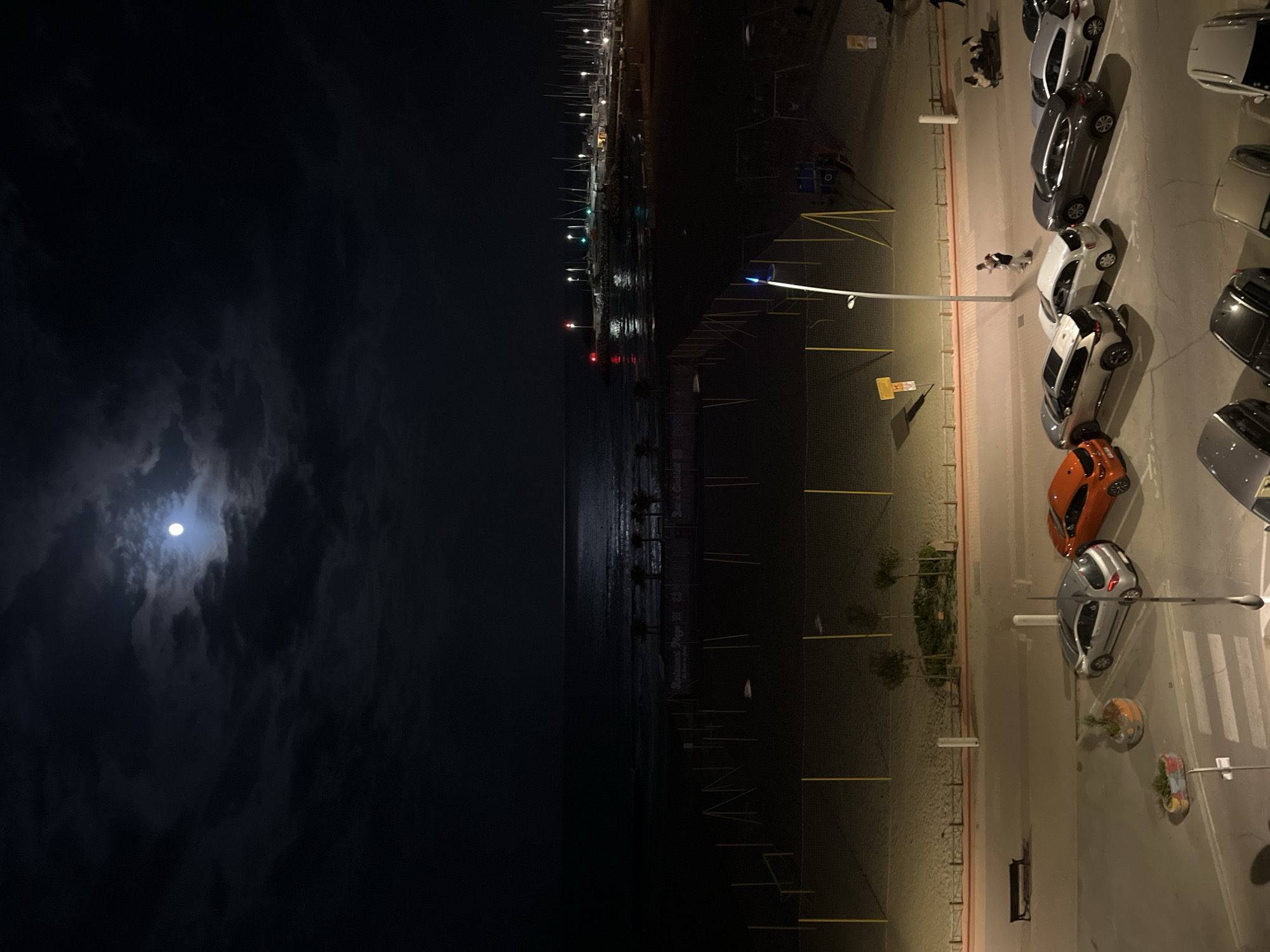 The image depicts a nighttime scene in Palavas-les-Flots, France, showcasing a coastal area under a clouded sky with a visible moon. To the left, the dark ocean meets a beach. A pier and moored boats are visible along the shoreline with lights reflecting in the water. The beach transitions into a lighted area with parked cars and street lamps on the right side of the image. Two figures are walking between the cars.