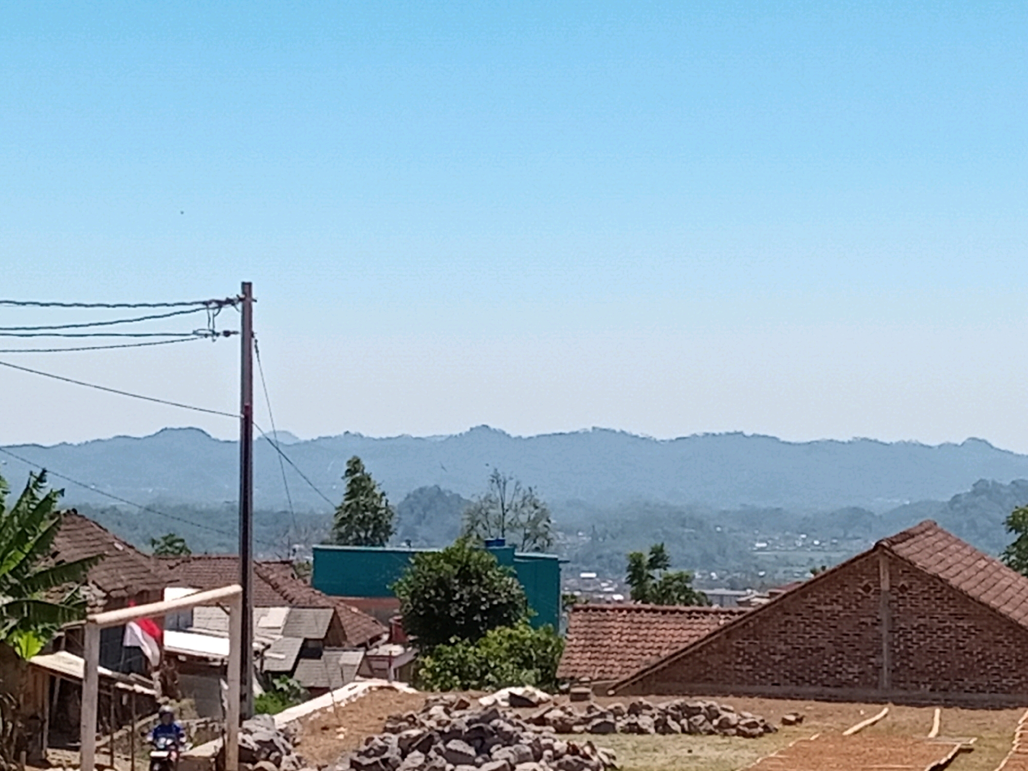 The image depicts a rural scene in Gandurejo, Indonesia. In the foreground, a pile of rocks sits on dry earth, with some lighter-colored wooden planks arranged on the ground, suggesting an outdoor drying area. A brick building with a tiled roof is prominent on the right. To the left, a cluster of houses with tiled roofs is visible, including one with a white and red Indonesian flag displayed. A utility pole with wires stands to the left of center. In the middle ground, a turquoise-colored building and trees are present. The background features rolling, mist-covered mountains under a clear blue sky. A person on a blue motorcycle is visible in the lower left quadrant of the image, near the houses.