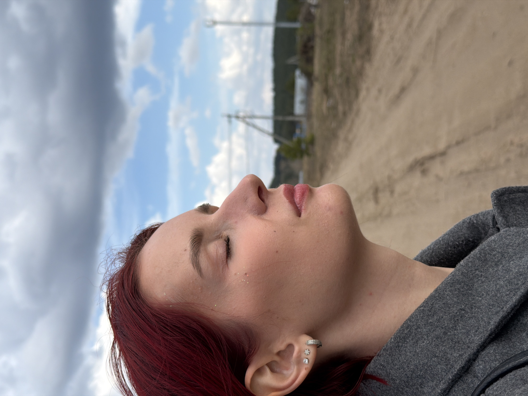 A close-up, profile shot captures a young woman outdoors, her head tilted upwards with eyes closed, seemingly in a moment of calm or contemplation. She is positioned on the right side of the frame, facing left. She has vibrant red hair, fair skin, and her lips are slightly parted. Her right ear is adorned with a silver hoop earring on the helix and two stud earrings on the lobe: one flower-shaped and one square-cut stone. Subtle glitter is visible on her cheekbone. She is wearing a grey textured coat or jacket.
The background is a blend of natural and semi-rural elements. The upper half features a dynamic sky, showing patches of bright blue with white, fluffy clouds interspersed with a significant mass of dark grey clouds, indicating varied weather. Below the horizon, a light brown dirt road or path stretches diagonally from the lower right towards the center, suggesting an open, unpaved area. In the middle distance, utility poles with wires are visible against the sky, backed by a blurry treeline, hinting at a rural or exurban environment in Chaikovsky, Russia. The lighting suggests it is daytime. There is no visible text in the image.