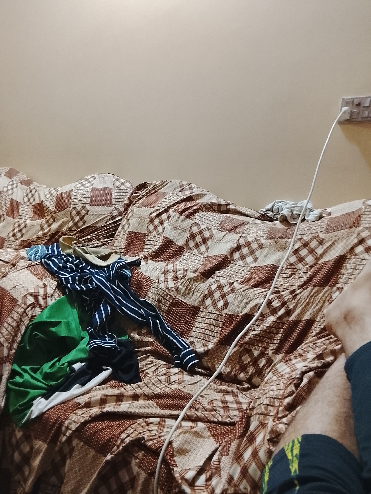 A cluttered bed with a brown and beige patterned bedspread dominates the scene. A pile of clothes, including items in green, black, white, and blue striped fabrics, is located towards the left side of the bed. A white charging cable runs from a power outlet on the wall, across the bed, and towards the lower right corner of the image where a person's legs are visible, clad in dark blue pants with a hint of yellow pattern near the hem. The walls are a light beige color. The location is presumed to be inside a residence in Multan, Pakistan, but this cannot be confirmed with certainty solely from the visual information. No specific actions or interactions are evident.