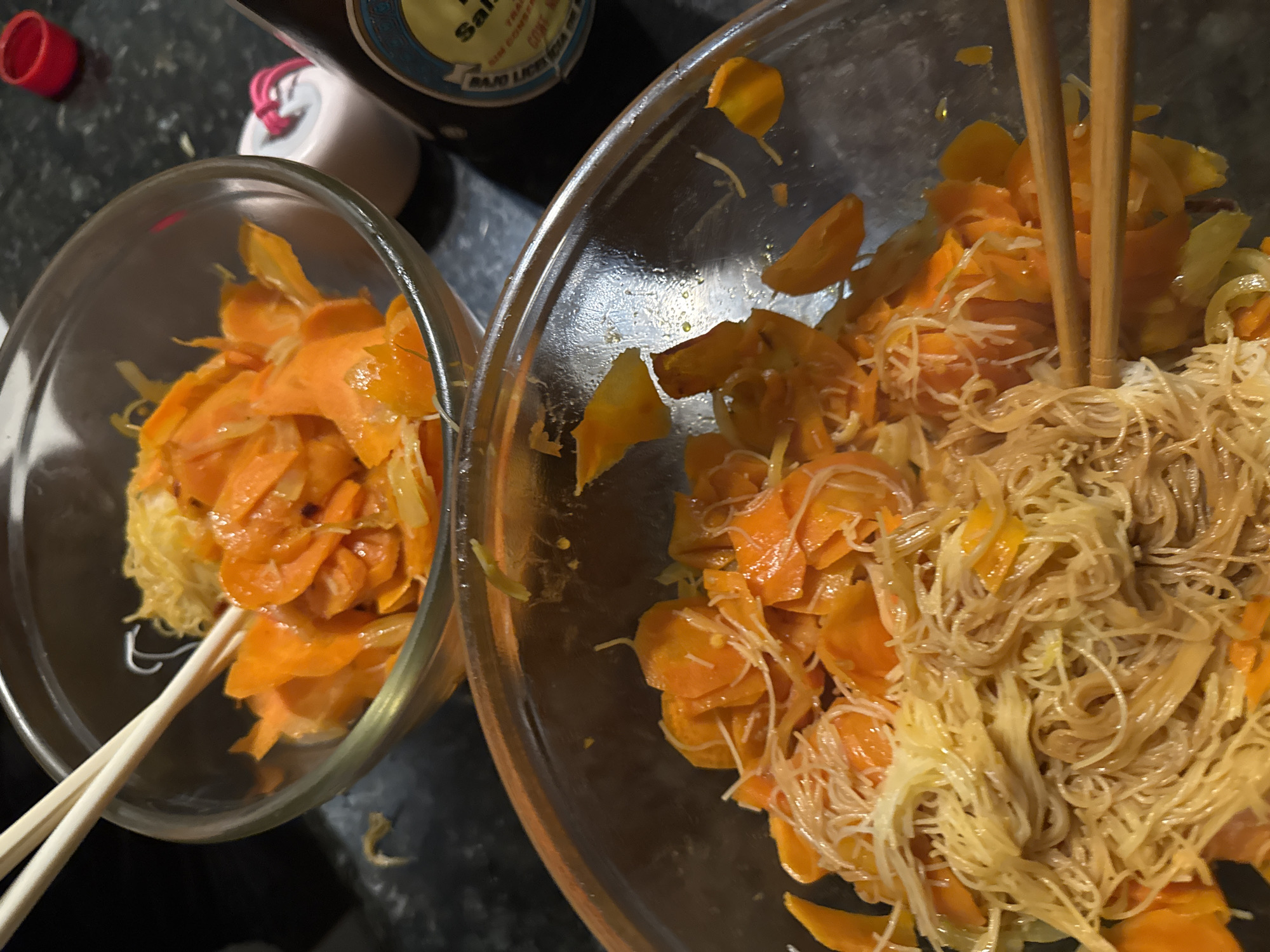 The image shows two clear glass bowls on a dark countertop. The bowl on the left is smaller and contains orange slices and a yellow, stringy substance, possibly egg noodles. Chopsticks are visible, partially submerged in the food. The bowl on the right is larger and contains the same orange slices and yellow noodles, with chopsticks also visible in the bowl. A bottle, partially visible at the top, suggests a sauce or dressing ingredient. The setting is likely a kitchen, based on the context of food preparation. Given the presence of the bottle, the location can be confirmed as Mexico City.