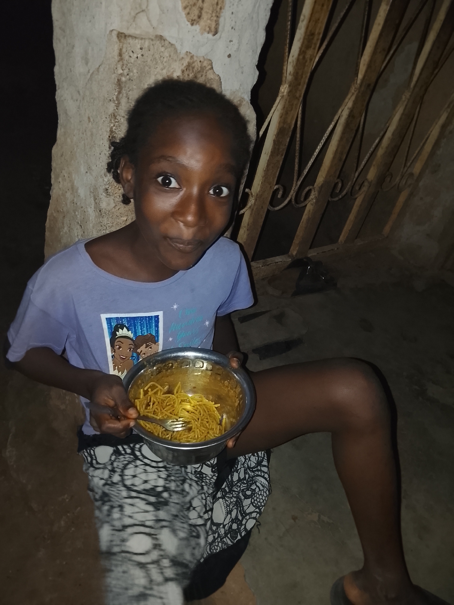 A young Black girl, appearing to be between 8 and 12 years old, is sitting on the ground in a dimly lit, rustic environment, likely in Akwanga, Nigeria. She has dark hair styled in braids or twists. She is wearing a light purple short-sleeved t-shirt with a graphic print featuring Disney's Princess Tiana and Prince Naveen. Visible text on her shirt includes "Always A Dream" and "Your Smile," surrounded by small stars. She is also wearing a black and white patterned skirt or wrap.
The girl holds a silver metallic bowl filled with yellow noodles, resembling spaghetti or instant noodles, with a silver fork resting inside. She looks directly at the camera with wide, expressive eyes and a slightly open mouth, conveying a sense of curiosity, surprise, or playful readiness.
The setting appears to be either outdoors at night or in a very dimly lit indoor space. Behind her, to the left, is a rough, unpainted light-colored concrete or stone wall. To her right and in the background, there is a rusty, decorative metal gate or grille, possibly part of a security feature for a door or window. The ground she sits on is dark, likely concrete or packed dirt. A pair of dark sandals or slip-ons are visible on the ground behind her. The scene captures a candid moment of a child about to enjoy a meal.