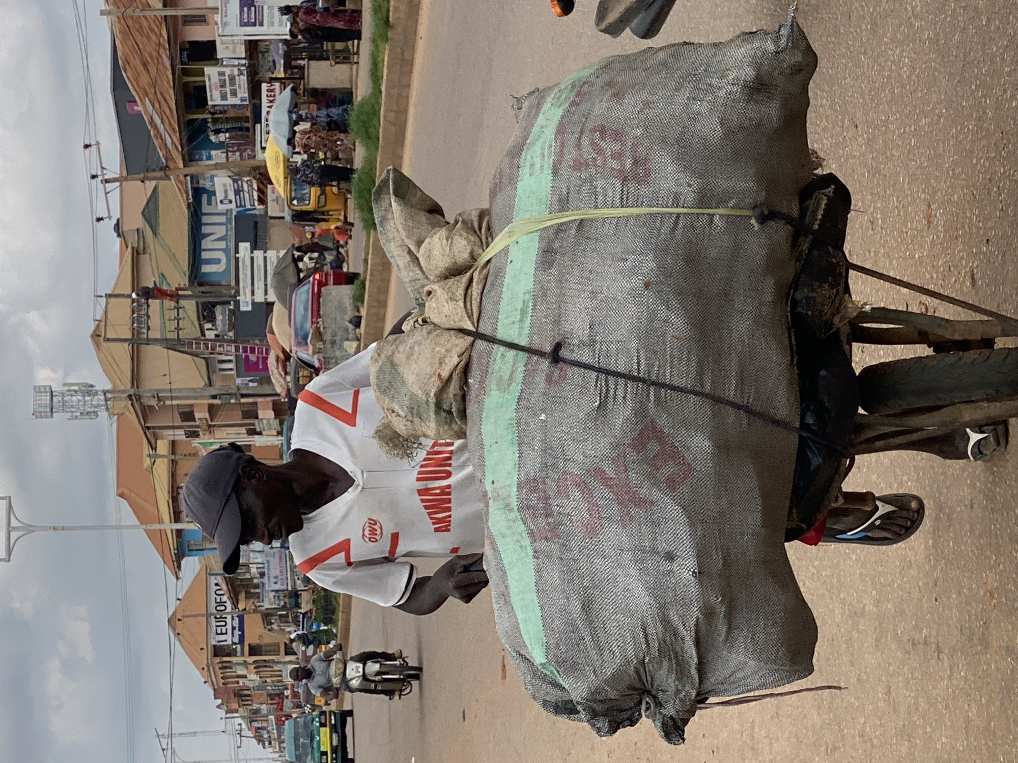 A man wearing a grey baseball cap and a white t-shirt with red trim, the words "OWU" and "AKWA UNITED" visible, pushes a cart loaded with two large, heavy sacks on a paved street in Sabon Tasha, Nigeria. The sacks are gray with a light green stripe across the top one. The man is in the foreground, in profile, facing right, and appears to be walking. The cart is laden and obscures much of his body from the waist down, though his left foot, wearing a flip-flop, is visible. In the background, the street is lined with buildings, some appearing to be shops or homes with signs, and other people are visible walking and on motorcycles. A yellow auto-rickshaw is also present in the mid-ground. The sky is overcast.