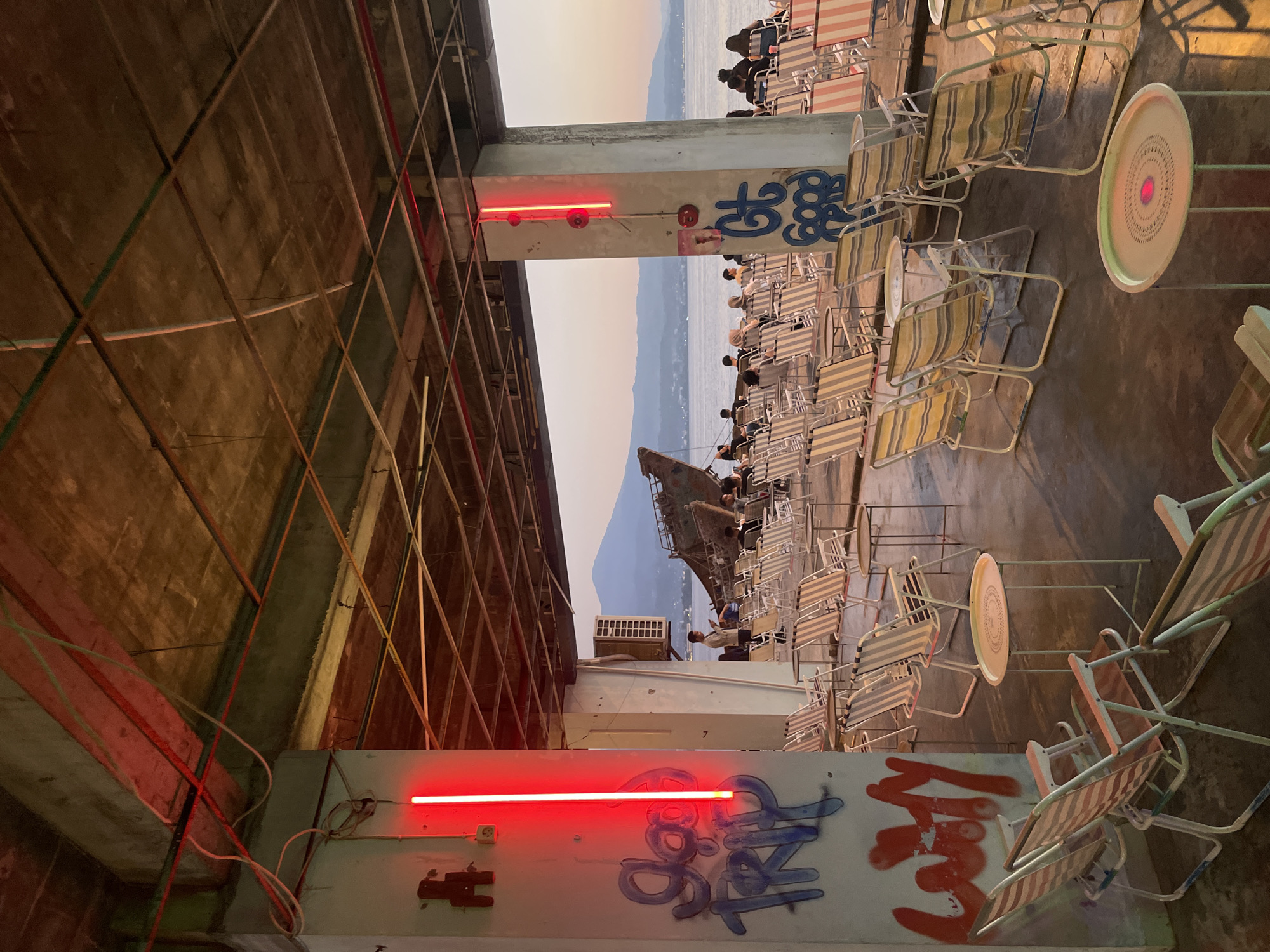 The image depicts an outdoor seating area with multiple rows of folding chairs and small tables, set under a concrete overhang with exposed beams and wiring. Red neon lights are mounted vertically on concrete pillars. Graffiti, including "go TRIP" and "lazy", is visible on the pillar in the foreground. In the background, a body of water stretches to the horizon, with mountains visible in the distance. A partially submerged shipwreck lies in the water. Several people are seated in the chairs, facing the water. The overall atmosphere suggests a casual, waterfront establishment. The scene is located at the "Kaki Langit Kopi" cafe in Manado, Indonesia.