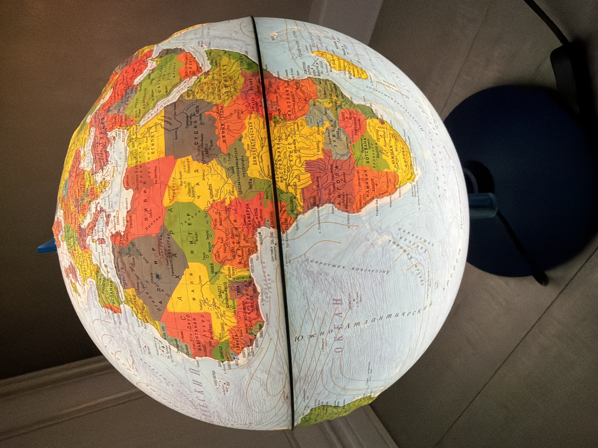 The image shows a globe depicting a map of the world. The continents of Africa and parts of Europe and Asia are visible. The globe is illuminated from within, causing the landmasses to stand out. The oceans are labeled in Cyrillic script, suggesting that the globe is designed for a Russian-speaking audience. A blue base is visible on the right side of the image, indicating that the globe is likely resting on a stand. The location is not discernable from the image.
