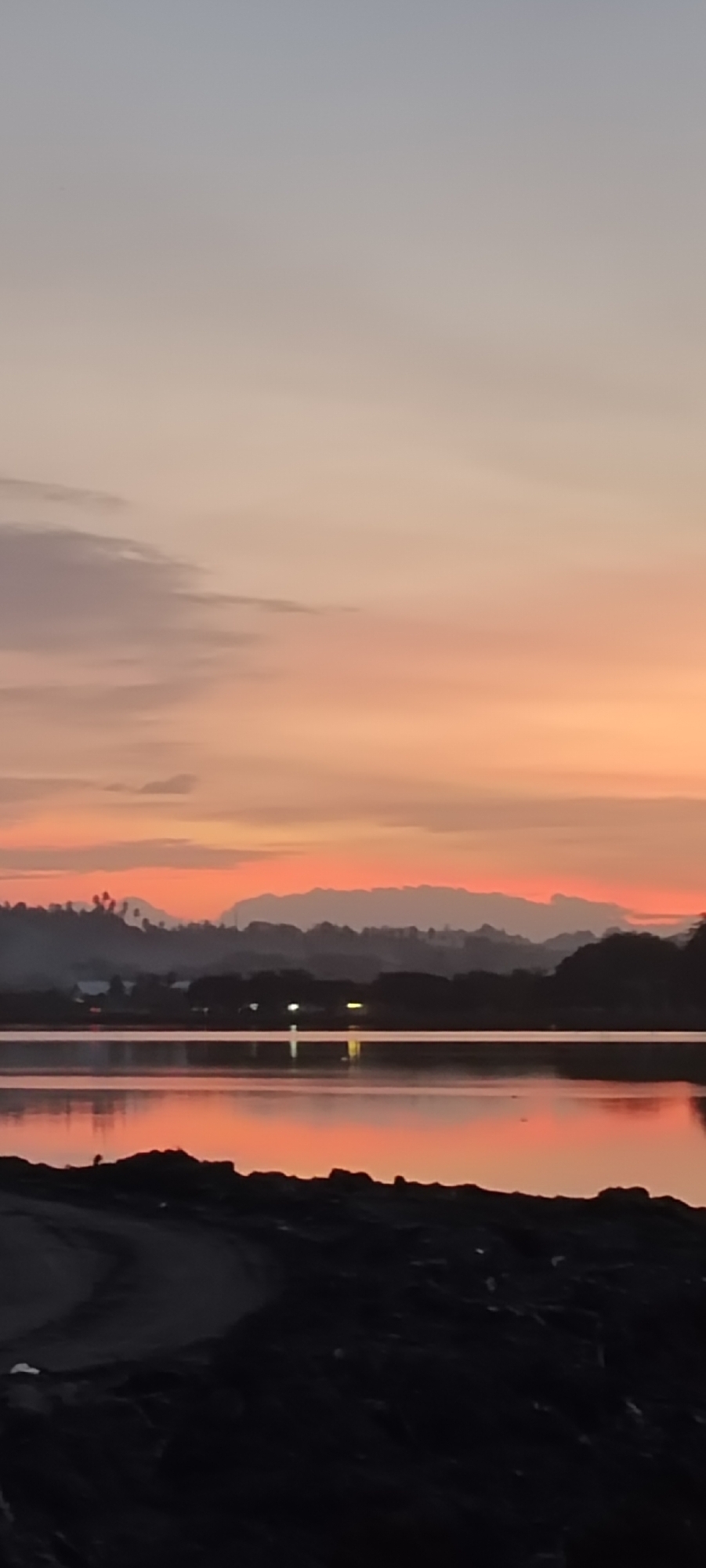 The image depicts a serene sunset over a body of water, likely the sea or a large lake, near the city of Lhokseumawe, Indonesia. The sky is painted with soft hues of orange, pink, and gray, with wispy clouds stretching across the horizon. In the mid-ground, a silhouetted treeline with scattered lights suggests a coastal settlement or a town situated near the water. Beyond that, a range of mountains forms a layered backdrop against the vibrant sky. The foreground features a dark, textured embankment, possibly a shoreline or a path made of rocks and debris. The water reflects the colors of the sunset, creating a warm and tranquil atmosphere. There are no people or explicit actions visible in the scene. The overall impression is one of natural beauty and peacefulness at dusk.