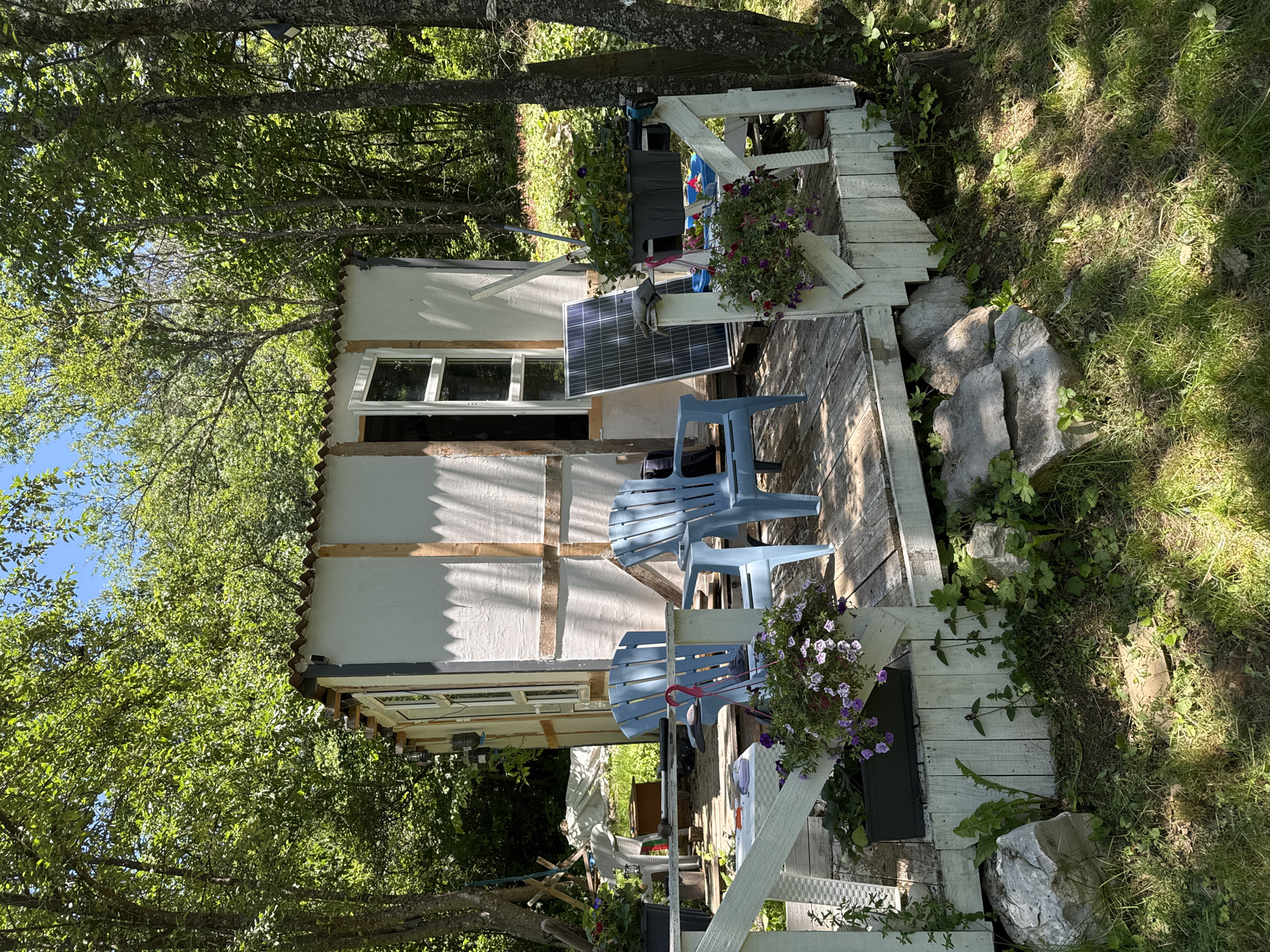 The image depicts an outdoor scene in Dramsha, Bulgaria, featuring a small, rustic structure situated in a wooded area. In the foreground, a weathered wooden deck is furnished with two light blue plastic chairs. Adjacent to the deck, a solar panel stands upright. The structure itself appears to be a simple, unfinished cabin with a corrugated roof and white walls framed by wooden planks. Green foliage from surrounding trees forms a canopy overhead, with sunlight filtering through the leaves. The ground is covered in grass and scattered rocks. The overall impression is one of a rural, DIY construction in a natural setting. There are no people visible in the image.